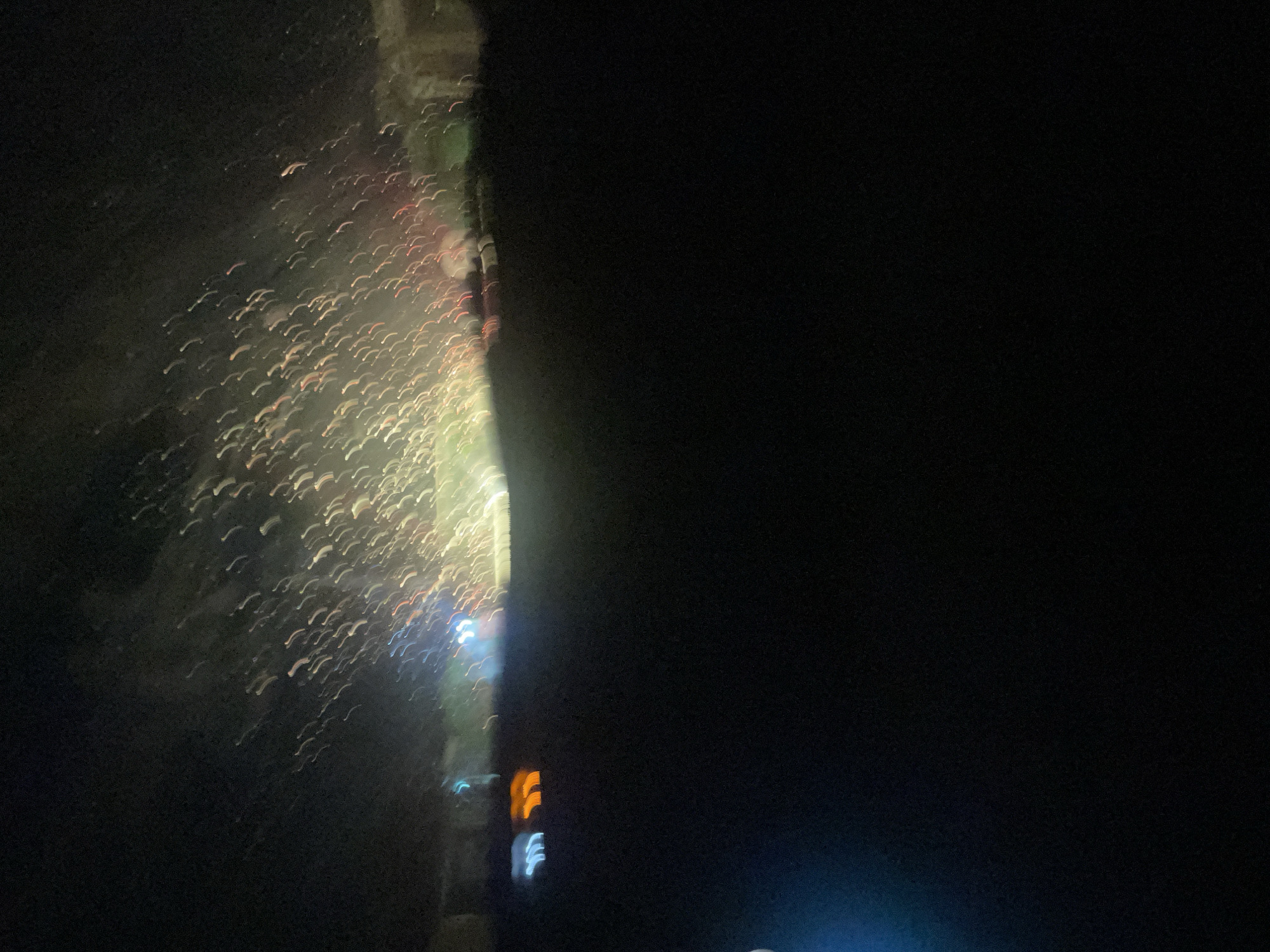 The image captures a dynamic nighttime scene in Ilorin, Nigeria, characterized by blurred streaks of light suggesting movement and activity. The left side of the frame is dominated by a vertical, blurry structure, possibly a building or a natural formation, illuminated by a cascade of light trails. These trails appear as numerous curved, elongated strokes of various colors, primarily white and yellow, with hints of red and blue interspersed. They suggest artificial lights, such as those from vehicles or perhaps even fireworks, creating a sense of energy and perhaps celebration.
The right side of the image is a vast expanse of deep blackness, indicating the absence of light or a very dark background, creating a stark contrast with the illuminated left side. Within this darkness, towards the bottom right, are faint, blurred shapes of colored lights. Notably, there are distinct orange and blue light forms, suggesting traffic lights or illuminated signs.
The overall impression is one of a vibrant, active urban environment at night, with the motion blur adding a sense of dynamism. The presence of artificial lights and the urban setting suggest the activity is taking place in a populated area. The time of day is clearly night, given the darkness. There is no discernible weather information, but the lights suggest it is not heavily obscured by fog or rain. No people or specific objects are clearly identifiable due to the motion blur and low light conditions. There is no visible text in the image.