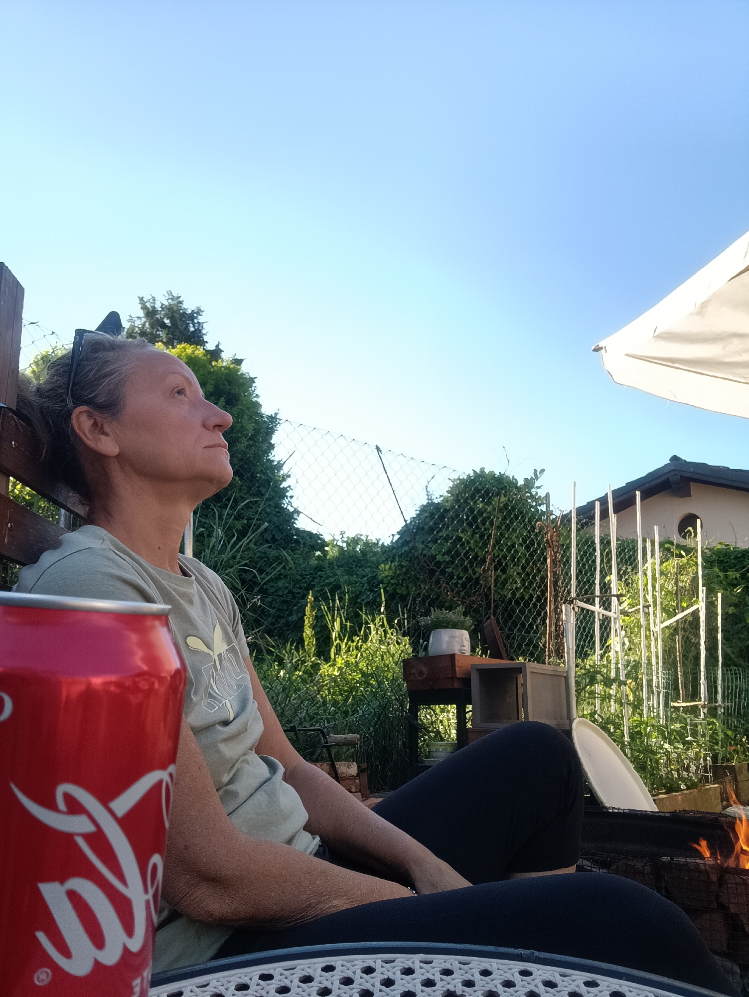 A medium shot captures a woman sitting outside in Viadana, Italy. She has sunglasses in her hair and is looking up at the sky. A red can of Coca-Cola is on a table in front of her. Behind her is a garden with a chain-link fence, greenery, and a small building. A fire is burning in the lower right corner of the frame. A white umbrella provides shade.