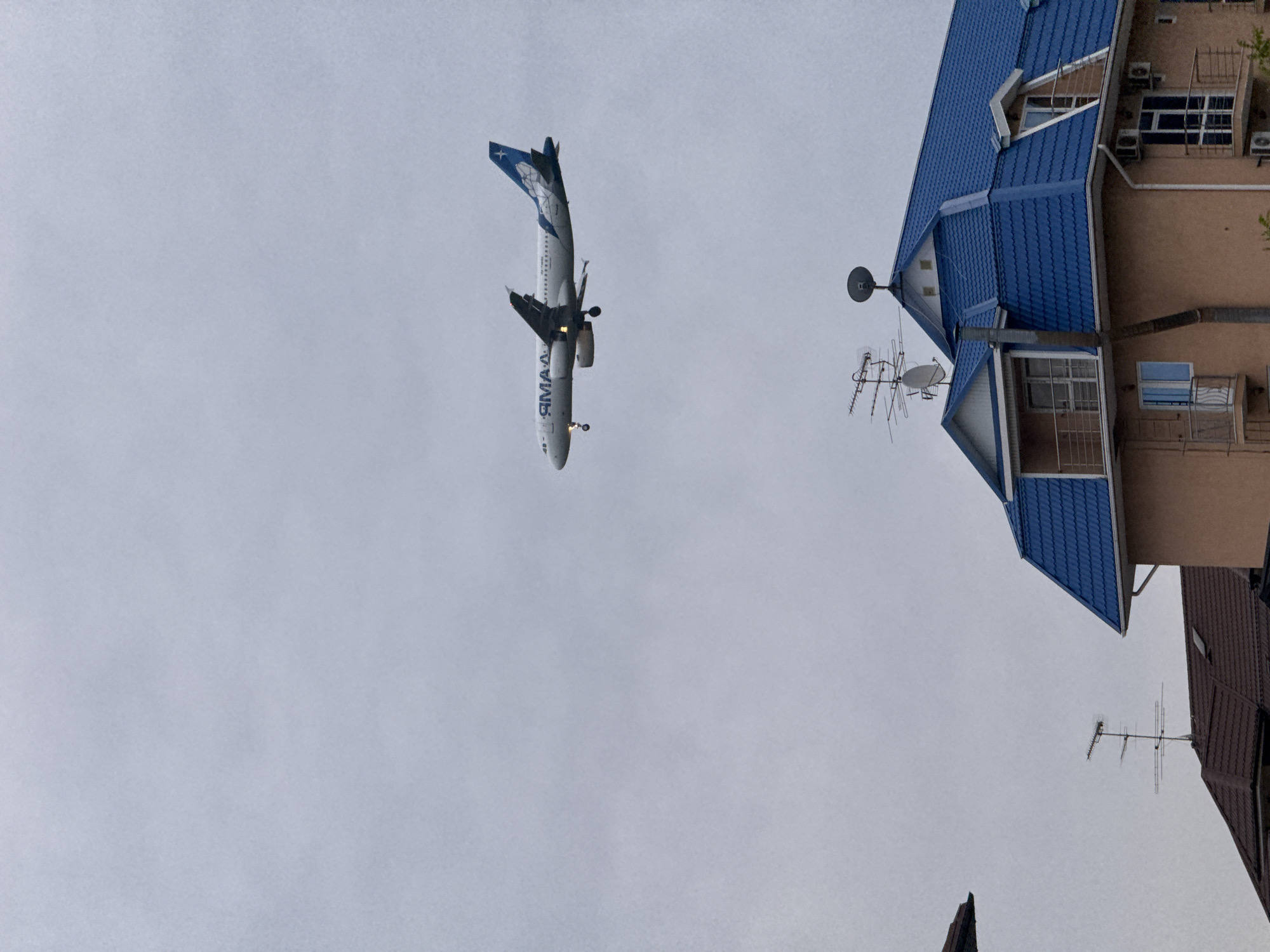 An aircraft, tail number VQ-BND (identified by "IRMAAA" on the fuselage), descends toward Sochi International Airport (AER) over a residential area in Sochi, Russia. The aircraft's landing gear is deployed. A blue-roofed building with satellite dishes and antennas is visible to the right. The sky is overcast.