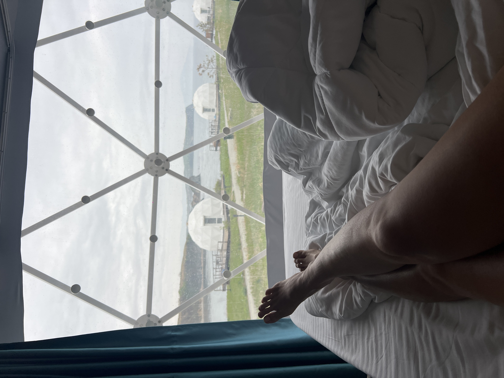 A person is lying in bed inside a dome-shaped structure. Outside the window, in Zarubino, Russia, are additional dome structures, a body of water, and green grass. The sky is overcast. The person's legs are visible from the thigh down to the toes, and they are under a white comforter and white sheets. A blue curtain partially covers the lower left corner of the window. The window is a geodesic dome structure with white beams and transparent panels.