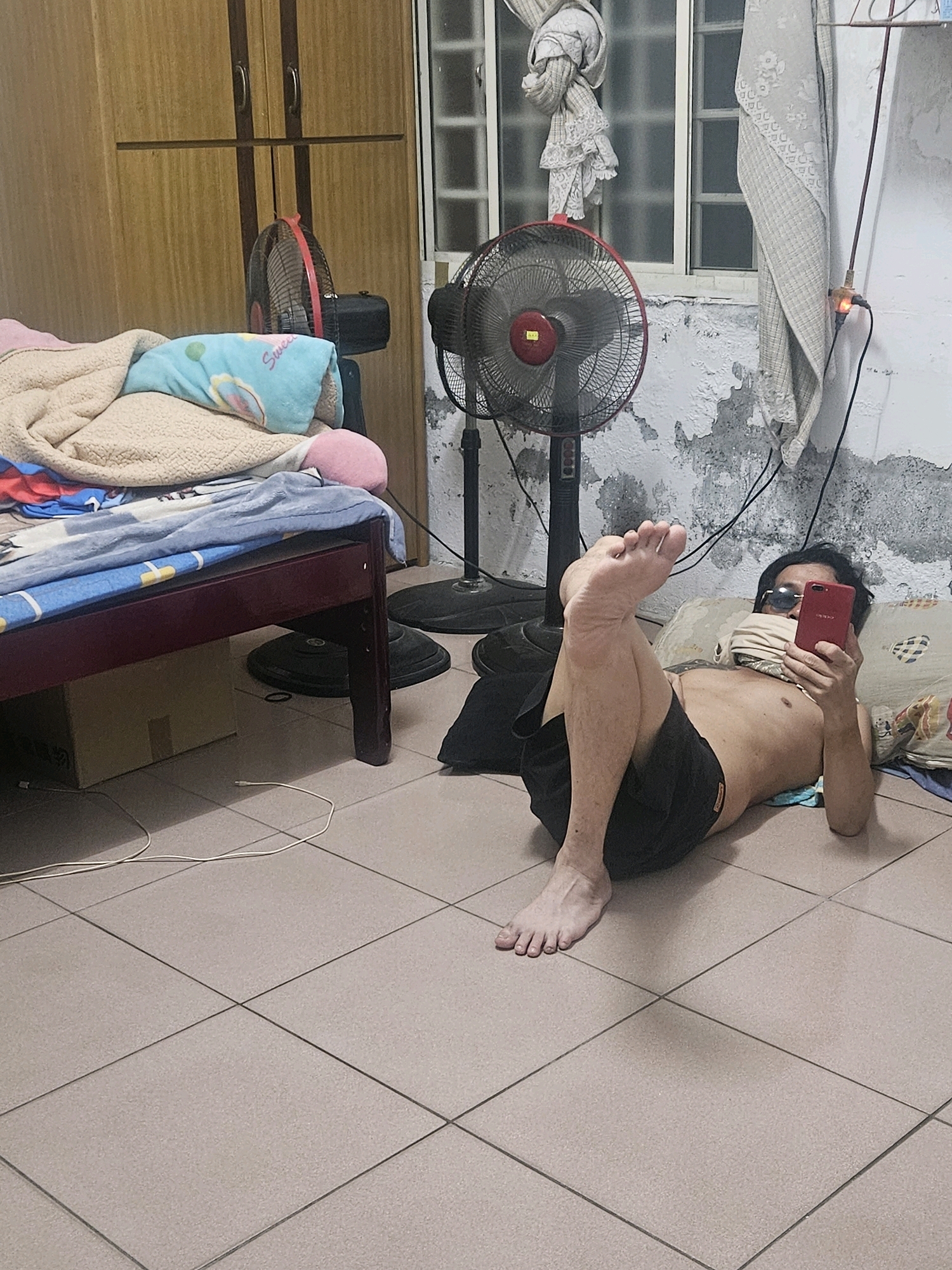 A man lies on the floor of a room, propped up on a pillow, holding a red smartphone in his right hand and taking a selfie. He is shirtless and wearing black shorts. His left leg is bent at the knee and extended upwards, with his foot resting on the floor. In the background, there is a bed with blankets and pillows, two electric fans, and a wooden wardrobe. A window with a curtain is visible on the wall. The floor is tiled. The overall environment suggests a domestic setting in Huwei Township, Taiwan.