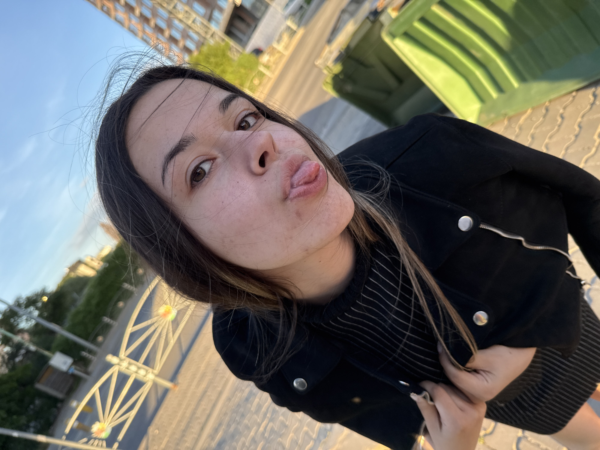 A young woman with long brown hair and wearing a black jacket and striped top is taking a selfie. She is sticking her tongue out and making a playful expression. The background includes a building with multiple windows, possibly an apartment complex. There is a green recycling bin on the right side of the frame. Additionally, the metal structure of a Ferris wheel can be seen on the left side of the frame. The location is likely in Sofia, Bulgaria, due to the specific type of Ferris wheel structure. The ground is paved with interlocking bricks. The overall lighting suggests it is late afternoon or early evening.