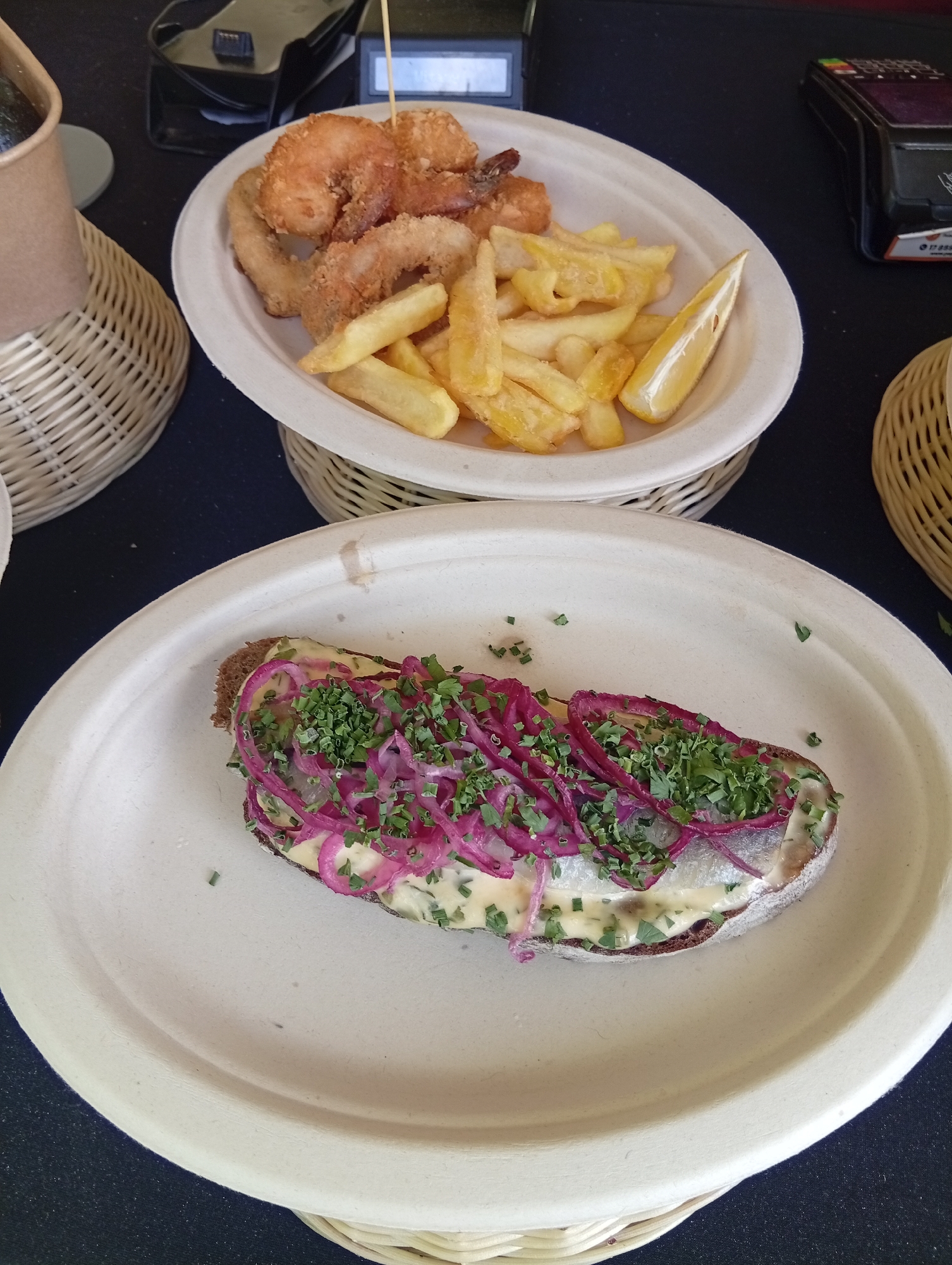The image captures a close-up, high-angle view of two dishes served on light-colored, circular, disposable plates. The top plate, positioned slightly behind and to the left of the bottom plate, contains a serving of fried seafood, likely shrimp, and french fries, accompanied by a wedge of lemon. The seafood pieces are golden brown and appear to be battered and deep-fried. The french fries are thick-cut and also golden. Both plates are presented within woven basket-like holders.
The bottom plate showcases an open-faced sandwich. A slice of dark bread serves as the base, topped with a creamy sauce, thinly sliced red onion rings, and chopped fresh green herbs, likely parsley or chives, and possibly some fish. The sandwich is arranged in the center of the plate.
In the background, a dark surface is visible, with elements of a transaction or service setup. To the left, a light-colored paper cup is partially visible, and behind it, a black electronic device, possibly a payment terminal or cash register, is present. A credit card reader is also visible on the right side of the frame. The overall presentation suggests a casual dining or street food setting. The lighting is natural and slightly diffuse, highlighting the textures and colors of the food. The scene is devoid of any identifiable landmarks or specific urban features that would pinpoint a location within Wrocław, Poland.