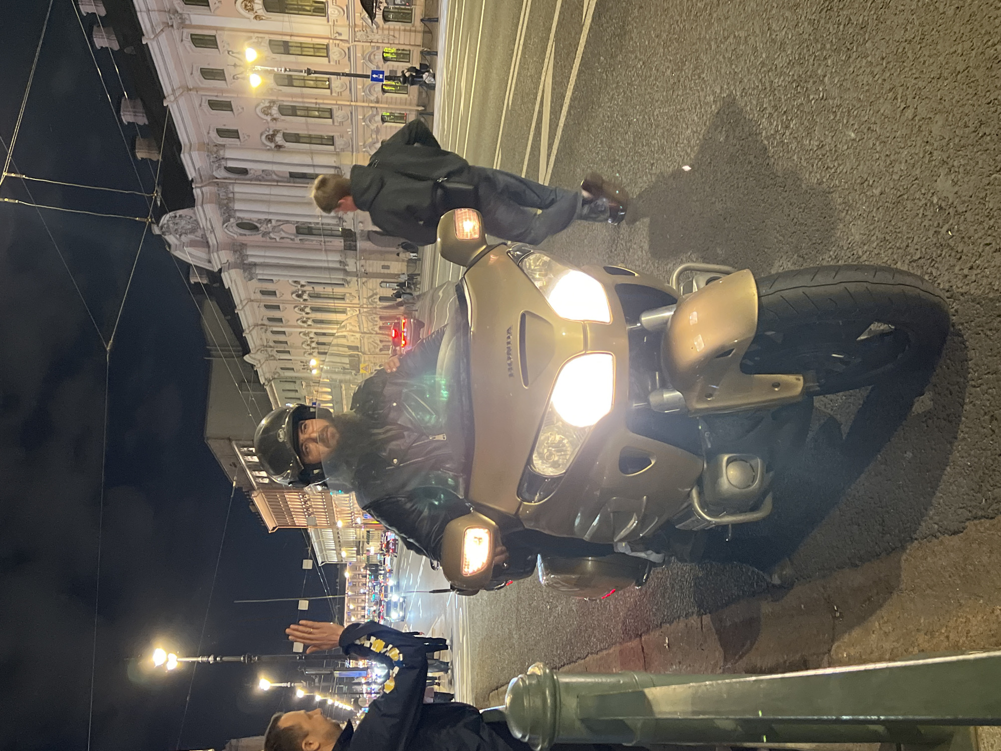 A nighttime street scene depicts a man on a gold Honda Goldwing motorcycle in what appears to be the vicinity of Palace Square, Saint Petersburg, Russia. The motorcycle is parked on the side of the road near a green iron railing, with the rider wearing a helmet and dark clothing looking at the camera. Near the motorcycle, there are two other men. One man in a black jacket and dark pants is bent forward, possibly walking. Another man is seen near the railing, facing away from the camera, with one hand raised. The buildings in the background are brightly illuminated, and the area seems to be a busy thoroughfare with streetlights and possible crosswalk markings on the road.