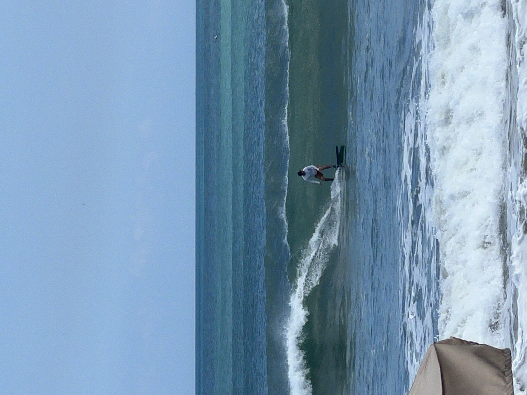A lone surfer rides a wave in the ocean off Kuta, Indonesia. The surfer is positioned in the middle of the frame, wearing a white long-sleeved shirt and orange shorts, and is standing on a black surfboard. The ocean displays multiple shades of blue and green, with waves breaking in the foreground and mid-ground. A light blue sky with faint clouds is visible in the upper portion of the image. In the lower left corner, a portion of a tan-colored beach umbrella is visible.