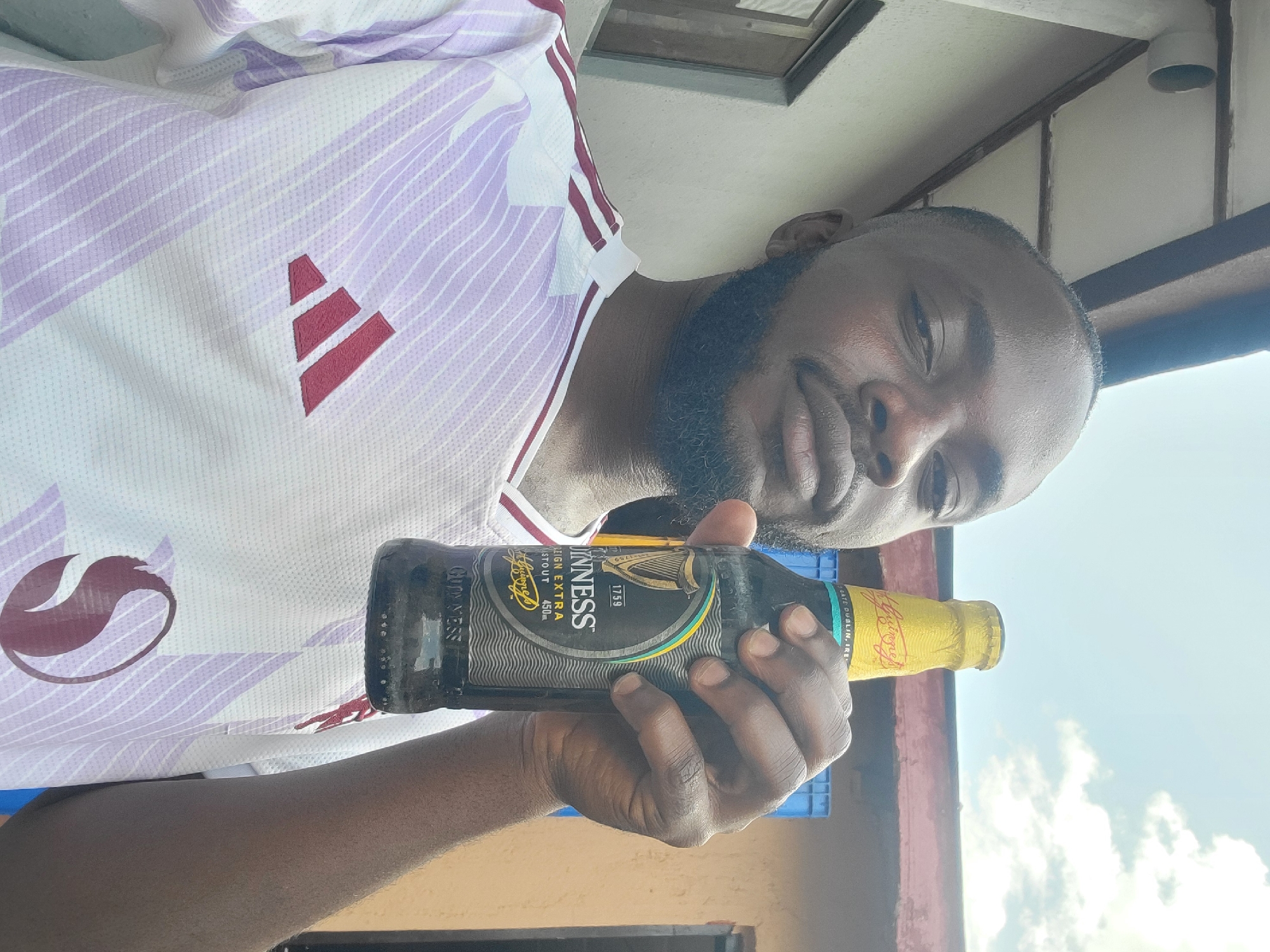 A man is taking a selfie outdoors, holding a bottle of Guinness Foreign Extra Stout. He is wearing a white and purple striped shirt with a maroon Adidas logo. The background suggests an outdoor setting with a building and sky visible. The scene is likely in Jos, Nigeria, as indicated by the provided context.