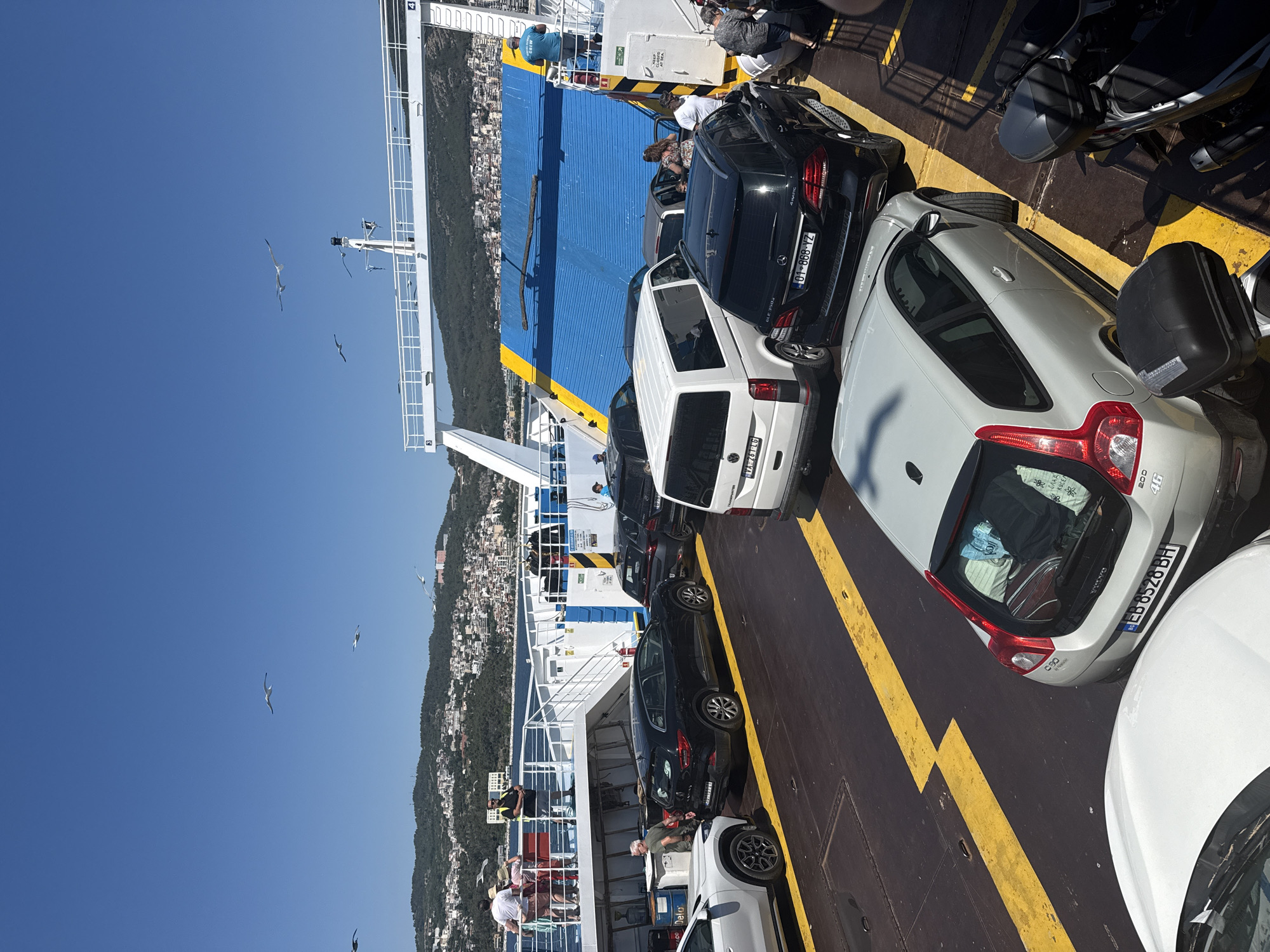 A high-angle, eye-level shot shows vehicles and people on a ferry. The ferry deck is black with yellow markings. A white car is in the foreground on the left, facing right. To its right is a silver Volvo C30, its rear hatch open, revealing luggage. Next to the Volvo is a black Mercedes-Benz SUV, and further right is a white panel van. Behind the van, a blue ramp extends upwards.
In the background, several people stand on an upper deck of the ferry, observing the scene. A coastal town with buildings and hills rises behind the ferry. The sky is a clear blue with a few seagulls flying. The location is identified as the port of Kavala, Greece.