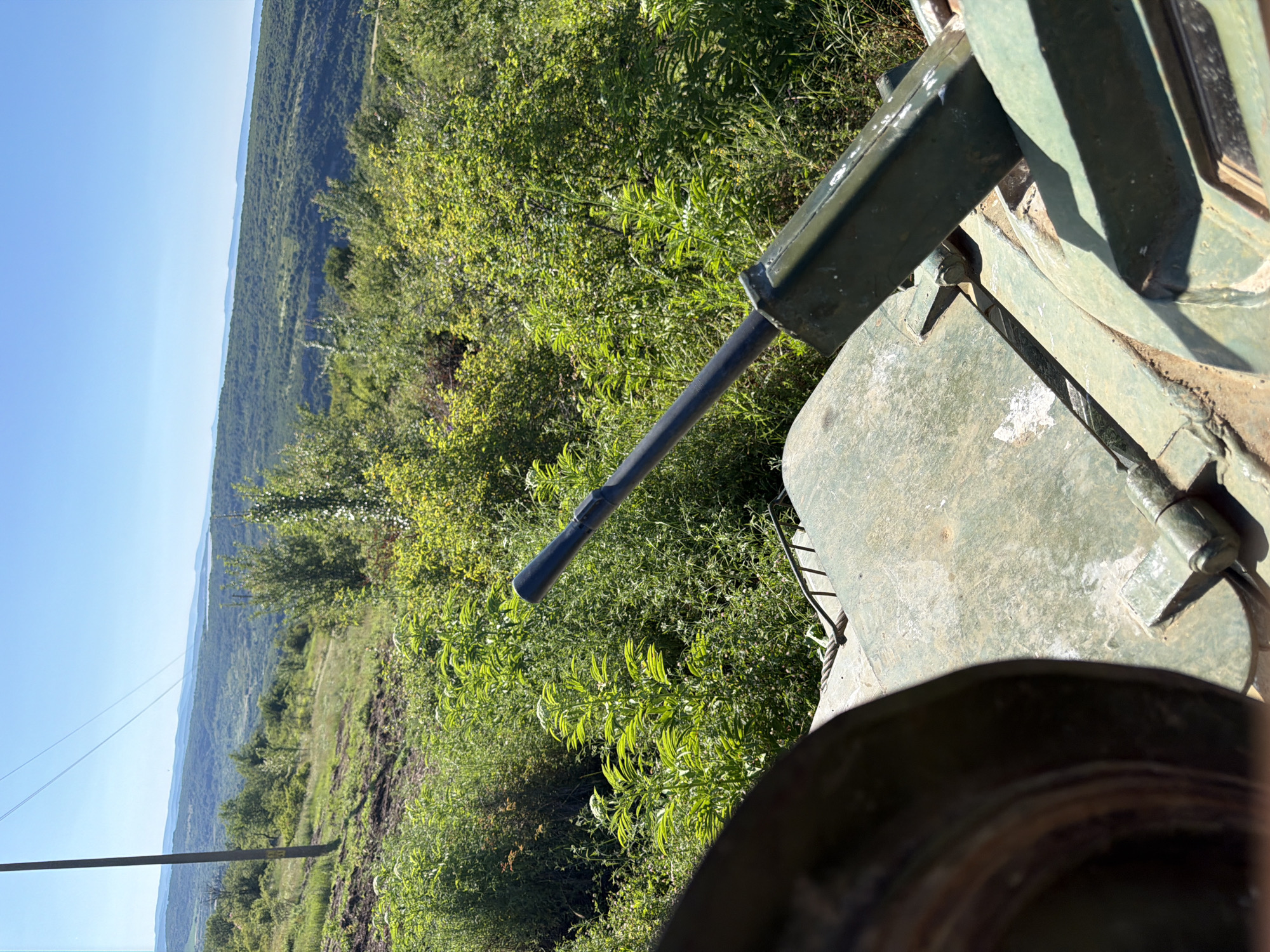 The image is a close-up shot of a green military vehicle, likely a tank, with its barrel pointing towards a lush, green landscape. The vehicle's exterior is weathered and shows signs of use. The barrel of the main gun is visible in the foreground, extending into the frame. The background consists of rolling hills covered in dense vegetation, with a clear blue sky above. A power line runs across the top of the frame, supported by a wooden pole on the left side. The scene is set in a rural, natural environment, with no people or specific structures visible apart from the military vehicle and the power pole. The location is identified as Beliakovets, Bulgaria.