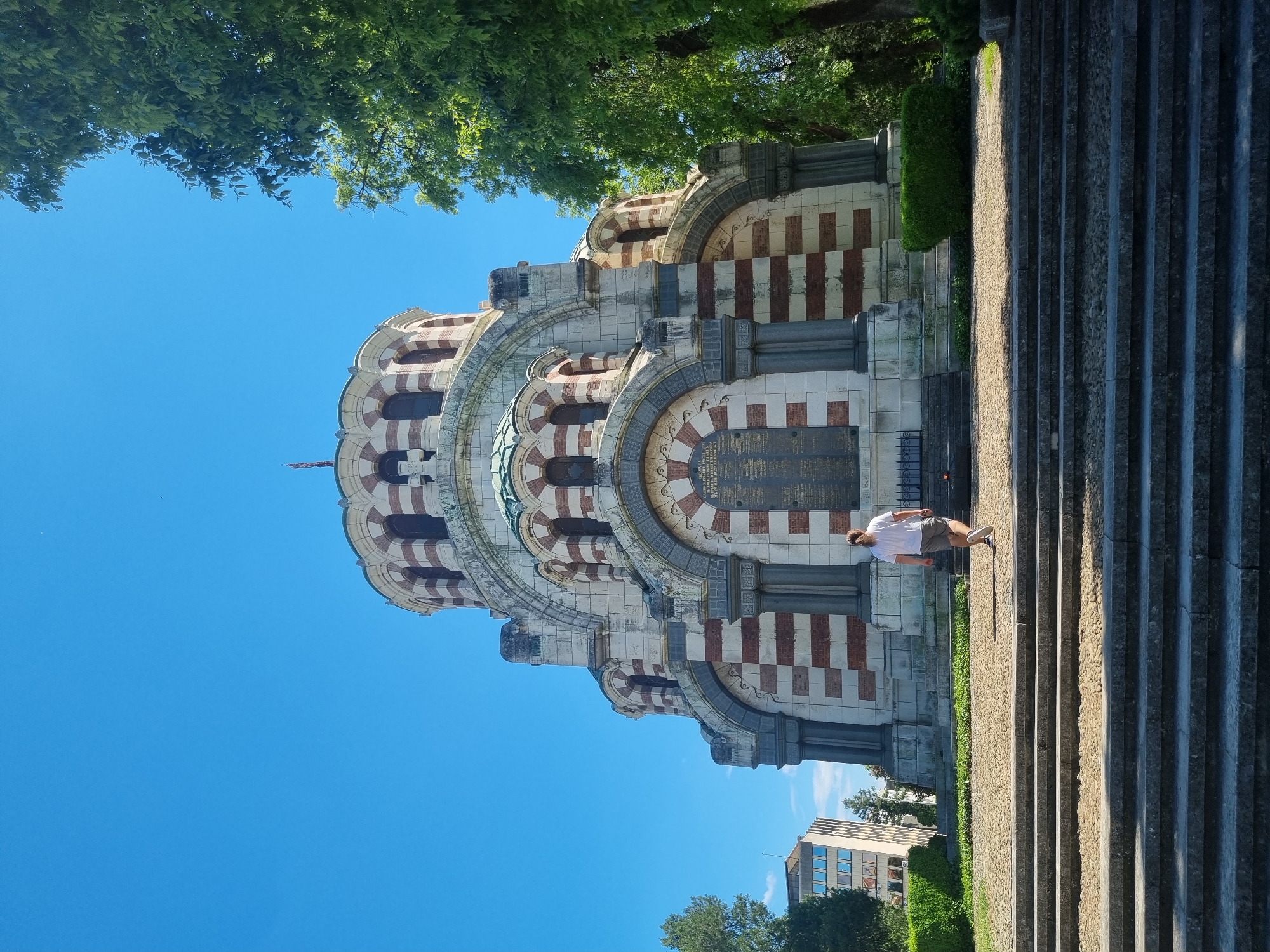 A vertical, eye-level shot captures the Saint George the Victorious Chapel Mausoleum in Pleven, Bulgaria, set against a clear blue sky and partially framed by green foliage. The mausoleum features a white stone facade with red brick detailing, a dome, and a commemorative plaque. A Caucasian male in a white shirt and shorts stands near the mausoleum. A concrete staircase extends into the foreground. A modern building is visible in the background, partially obscured by trees. The scene is brightly lit, suggesting a sunny day.