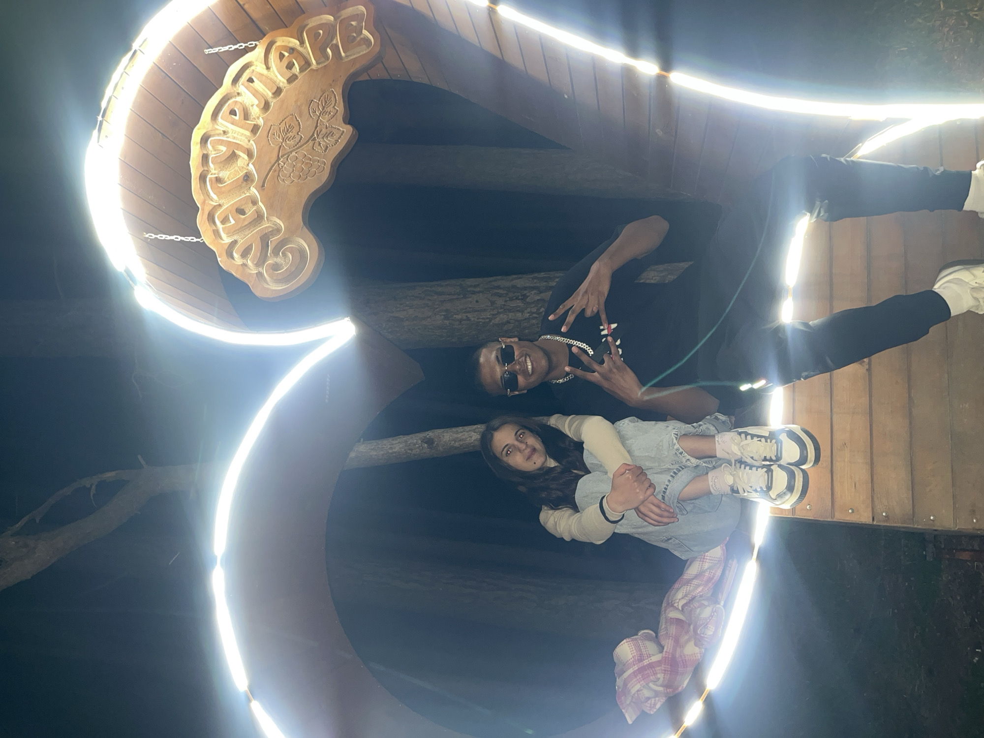 The image shows a low-angle shot of two people standing inside a lighted wooden structure in Sungurlare, Bulgaria. The structure is marked with "СУНГУРЛАРЕ," indicating the city name. One individual, a dark-skinned man, is positioned at the top of the frame, wearing sunglasses, a black T-shirt, and a chain necklace. He is making a "peace sign" with both hands. Below him, a light-skinned woman is sitting with her legs drawn up, wearing a light-colored long-sleeved shirt and denim shorts. A plaid blanket is draped over her legs. The structure appears to be a decorative arch or entrance, illuminated with bright lights around its edges, creating a tunnel effect in the photograph. The background is dark, suggesting an outdoor nighttime setting. The wooden planks of the structure are visible, and there are tree branches overhead.