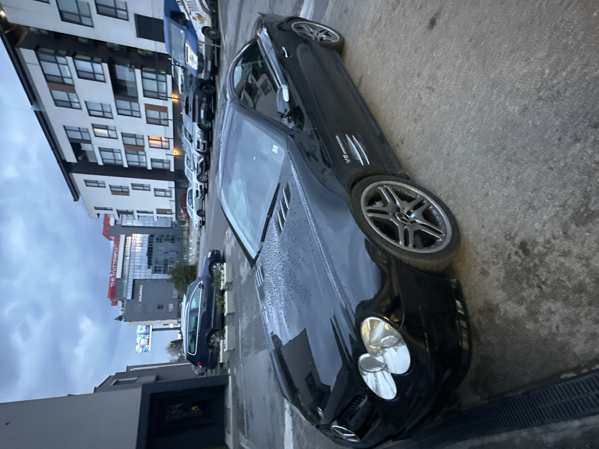 The image shows a black Mercedes-Benz SL parked on a street lined with other cars. The car is a V8 Kompressor model. In the background, there's a modern building with several floors and many windows. Further back, there's a building with the words "THE BUTTERFLY" and "GOLDWELL" visible on it. The scene appears to be located in Sofia, Bulgaria. The weather seems cloudy, as suggested by the sky's appearance.