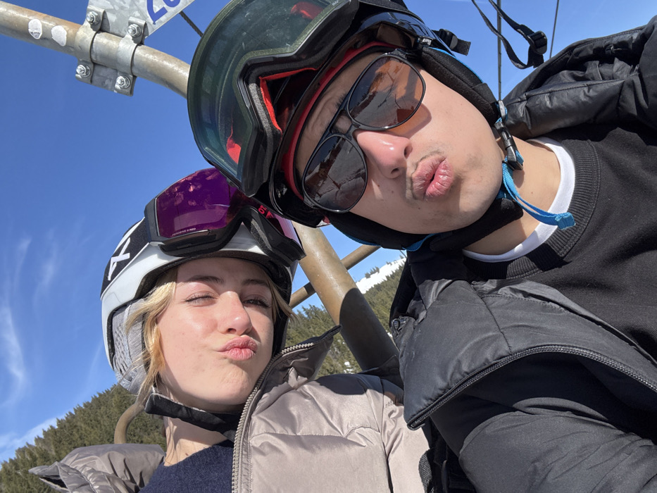 Two individuals are depicted on a ski lift in Manigod, France, under a clear blue sky. The person on the left, a female, wears a white and grey ski helmet with purple-lensed goggles attached, a dark top, and a light brown padded jacket. Her eyes are closed, and her lips are pursed. The person on the right, a male, wears a black ski helmet, dark aviator-style sunglasses, and ski goggles pushed up on his helmet. He has a mustache and is dressed in a black padded jacket over a black sweatshirt and a white t-shirt. His lips are also pursed. Both subjects are looking towards the camera. A metallic bar of the ski lift is visible behind them, along with a vertical metal pole with bolted attachments on the far right. The background features snow-covered terrain and evergreen trees under bright sunlight.