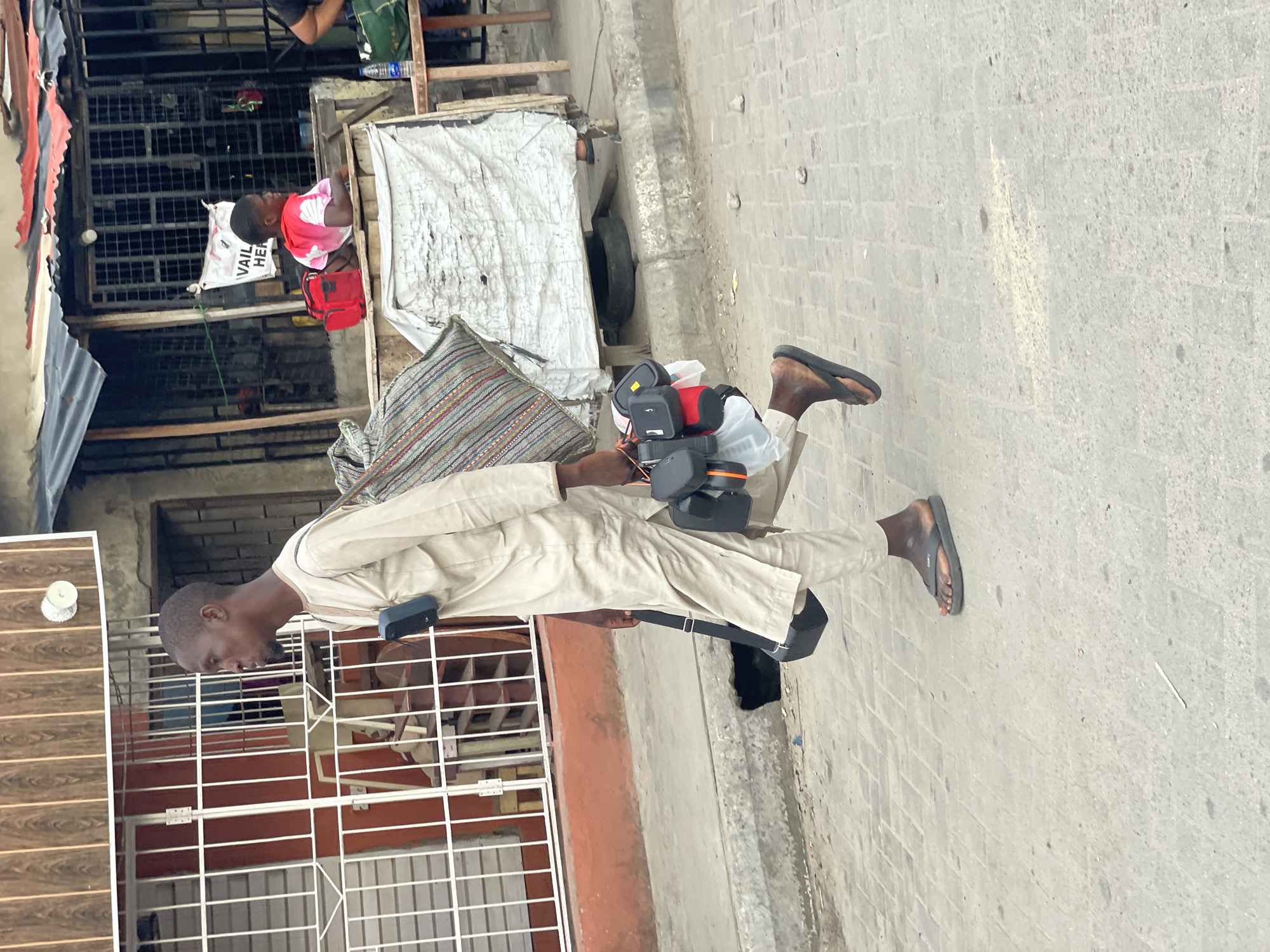 This outdoor photograph captures a street scene, rotated 90 degrees clockwise, likely in Ado, Nigeria, during the daytime with bright, clear weather.
The main subject is a man of dark complexion, wearing a light-colored (beige or khaki) long-sleeved tunic and black flip-flops. He walks on a paved sidewalk, his gaze directed forward. He is carrying multiple items, including a black, rectangular device strapped to his left hip and another attached to his chest/shoulder area. His right arm holds a pole from which several black and orange electronic devices (possibly speakers, radios, or power banks) are suspended in a clear plastic bag. He appears to be a street vendor or hawker, displaying his goods.
In the background, on the left side of the frame, there are several makeshift structures or stalls. A person, possibly a child, wearing a pink and white top, is seated behind one of these stalls, looking towards the right. Nearby, a red bag and a white bag are visible. The white bag has partially discernible text that reads "VAIL HERE," likely part of "AVAILABLE HERE." Further in the background, a person's upper torso and arm in a dark shirt are seen. The structures are characterized by corrugated metal, wooden planks, and metal grilles or gates. The sidewalk is made of interlocking paving stones, with some debris visible.