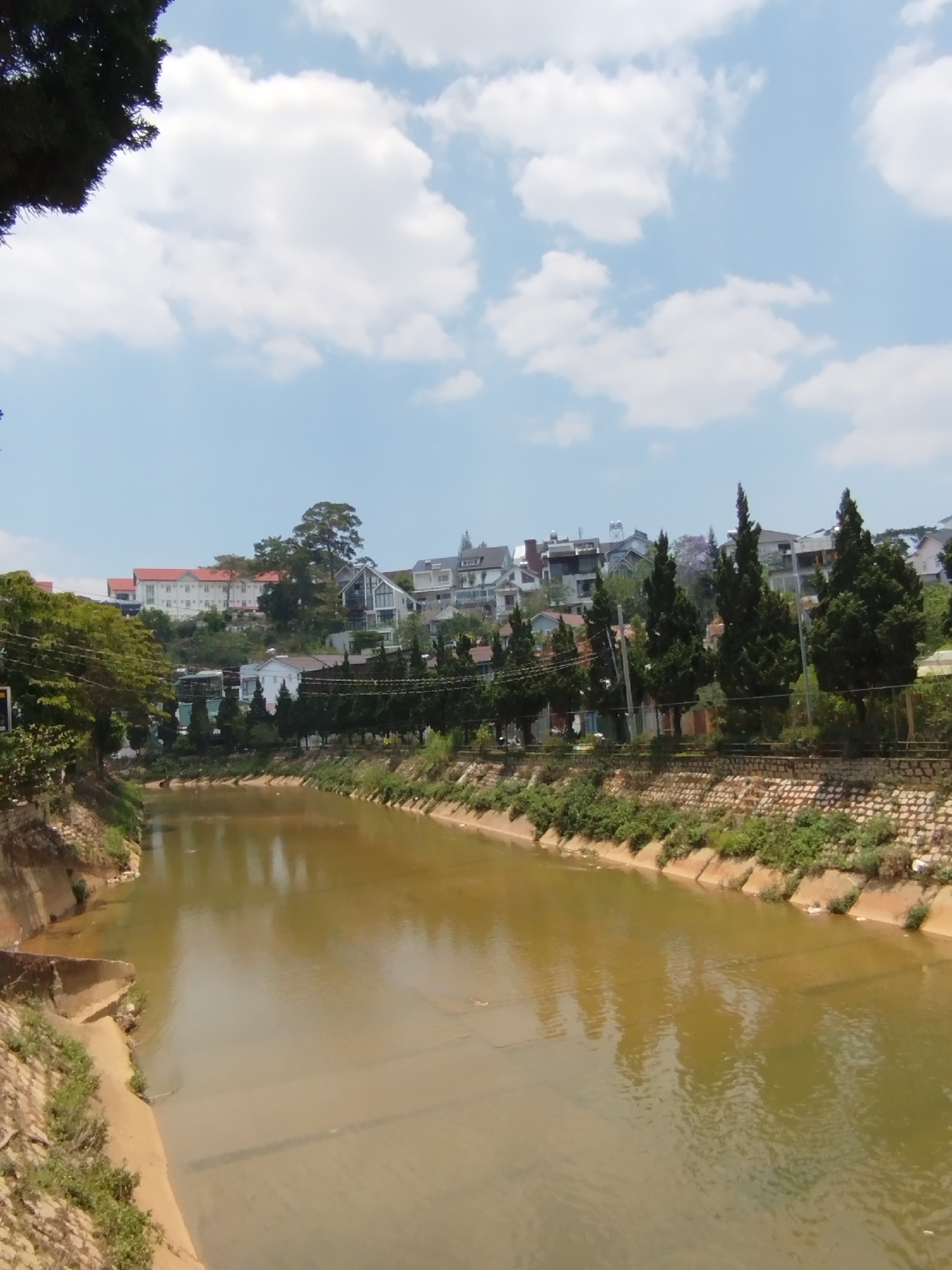 The image depicts a canal with murky water in the city of Cam Ly, Da Lat, Vietnam. The canal is lined with concrete banks and topped with stone walls and greenery. Houses and trees are visible on the hillside above the canal. The sky is blue with scattered white clouds.