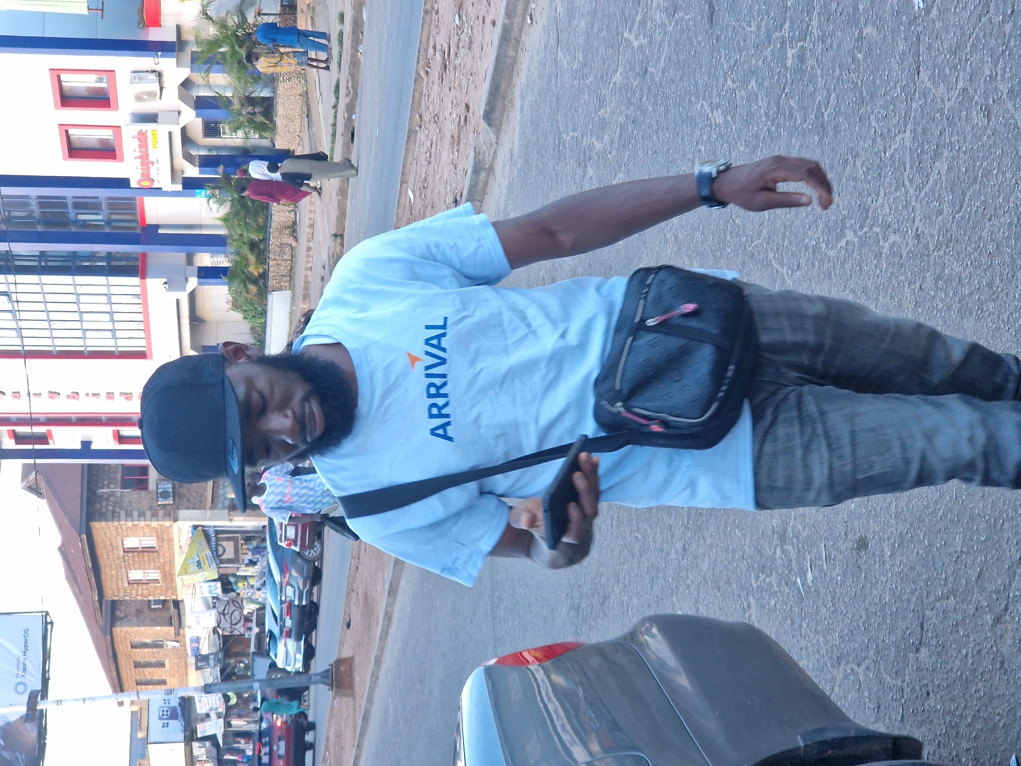 A man wearing a black baseball cap, a light blue t-shirt with "ARRIVAL" printed on it, and grey jeans is walking on a street in Jos, Nigeria. He is carrying a black crossbody bag and holding a black smartphone in his left hand. In the background, there are buildings, cars parked on the side of the road, and other pedestrians. The scene appears to be a busy street in an urban area.