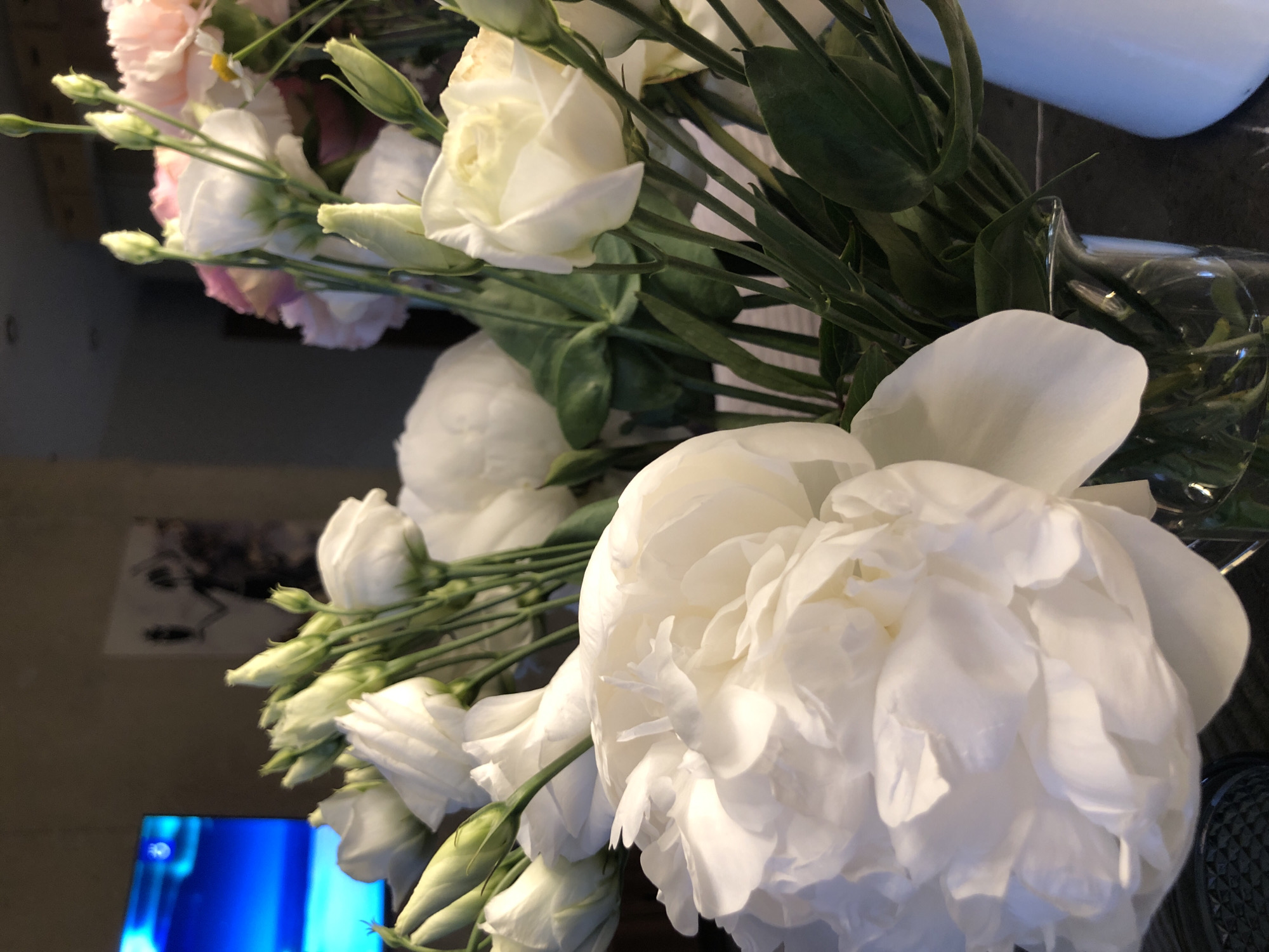 A close-up, eye-level shot captures a bouquet of white and pink flowers in a clear glass vase. The bouquet is dominated by large, ruffled white peonies in the foreground and mid-ground, with smaller white and pink lisianthus buds and blooms interspersed throughout. Dark green leaves and stems add depth and contrast. The background is softly blurred, with hints of a television screen displaying a blue image on the left and a framed artwork on the wall behind the flowers. The scene appears to be indoors, possibly in a domestic setting, with a dark countertop visible in the lower portion of the frame. The lighting is soft, highlighting the delicate textures of the flowers. The location is not discernible from the image.