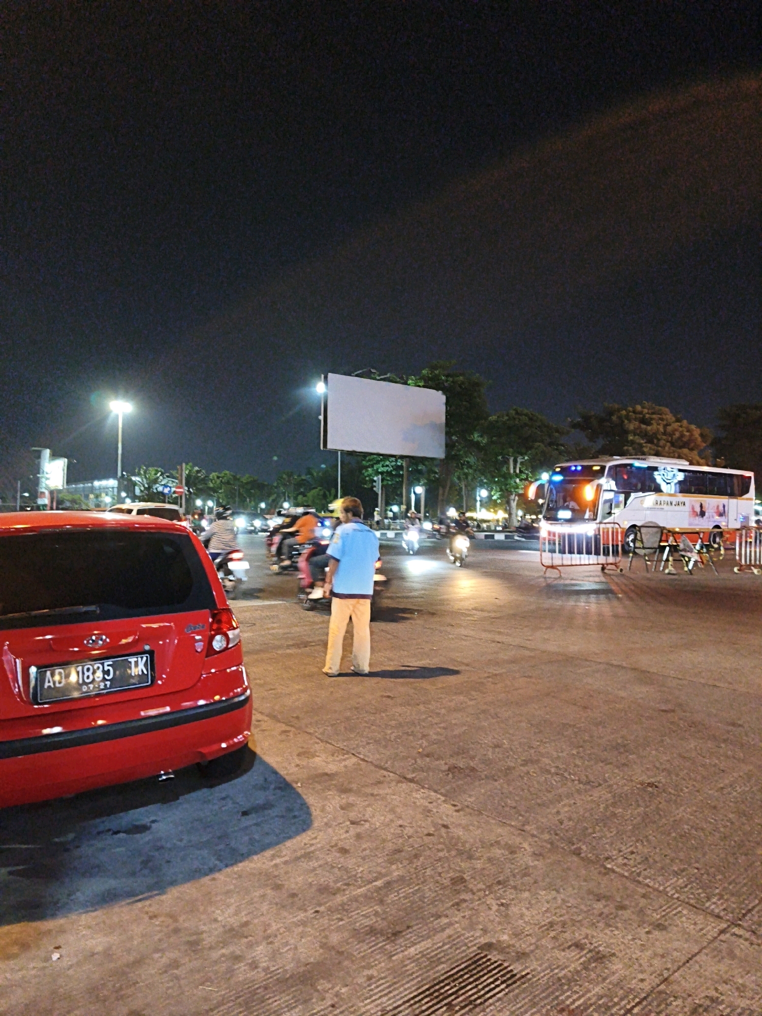 The image depicts a nighttime street scene in Surakarta, Indonesia, with a focus on a red car parked on the right side of the frame. The car's license plate reads "AD 1835 TK". A person wearing a blue shirt and orange pants stands in the middle of the road, facing away from the camera.
In the background, several motorcycles are visible, some with riders. A large white bus with the words "HARAPAN JAYA" on its side is parked to the right of the person in the blue shirt. The bus has its headlights and interior lights on. There are also trees and streetlights in the background, as well as a large, blank billboard. The road surface is paved with concrete and appears somewhat wet in places. The overall atmosphere is that of a busy street at night.