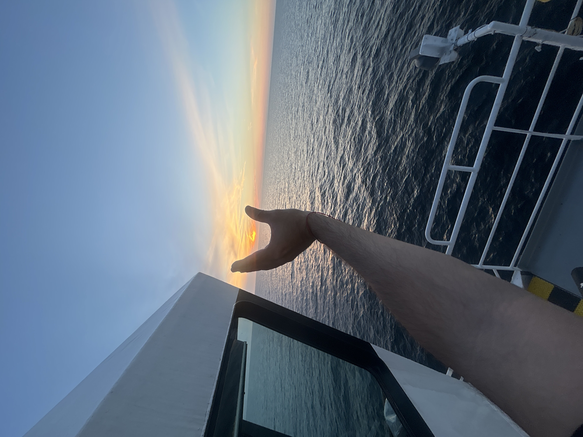 A first-person perspective shot shows a person's arm and hand holding the setting sun over the ocean. The hand is held up in front of the person, with the thumb and index finger forming a "C" shape around the sun. The sun is low on the horizon, casting a warm glow across the water and the sky. The ocean is calm, with gentle waves. The sky is a clear blue, with a few wispy clouds. In the foreground, the edge of a boat is visible, with a white railing and part of a cabin. The boat appears to be moving, as evidenced by the slight blur of the water. The scene is peaceful and serene.