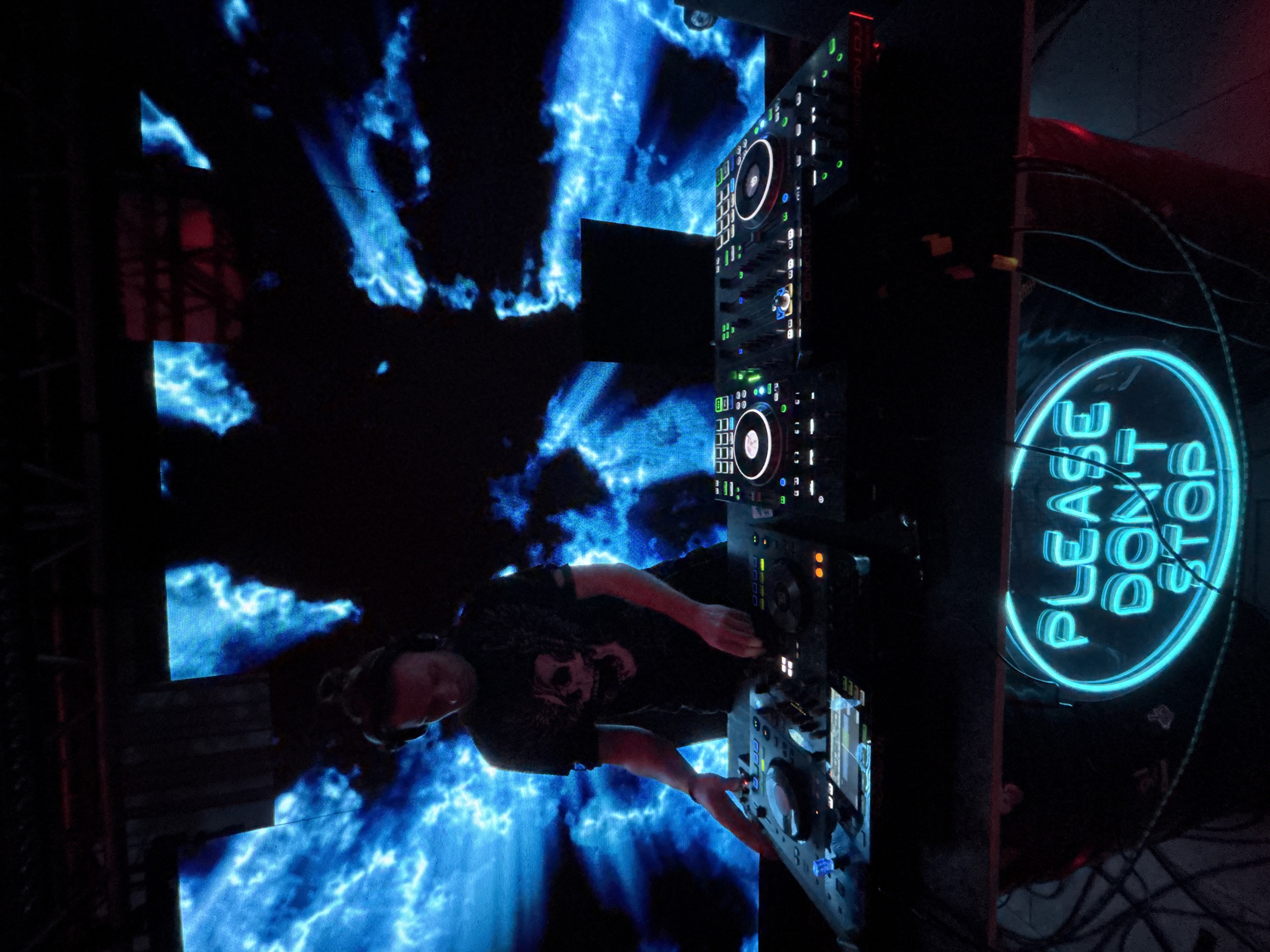 The image shows a DJ performing at a nightclub. He stands behind a DJ booth with a Denon DJ setup, including CDJs and a mixer. The background features a large LED screen displaying abstract blue visuals. A neon sign reading "PLEASE DON'T STOP" is visible to the right. The location is likely a nightclub, but the exact location in Votkinsk, Russia, is not ascertainable from the image. The DJ is wearing headphones and a t-shirt with a skull design.