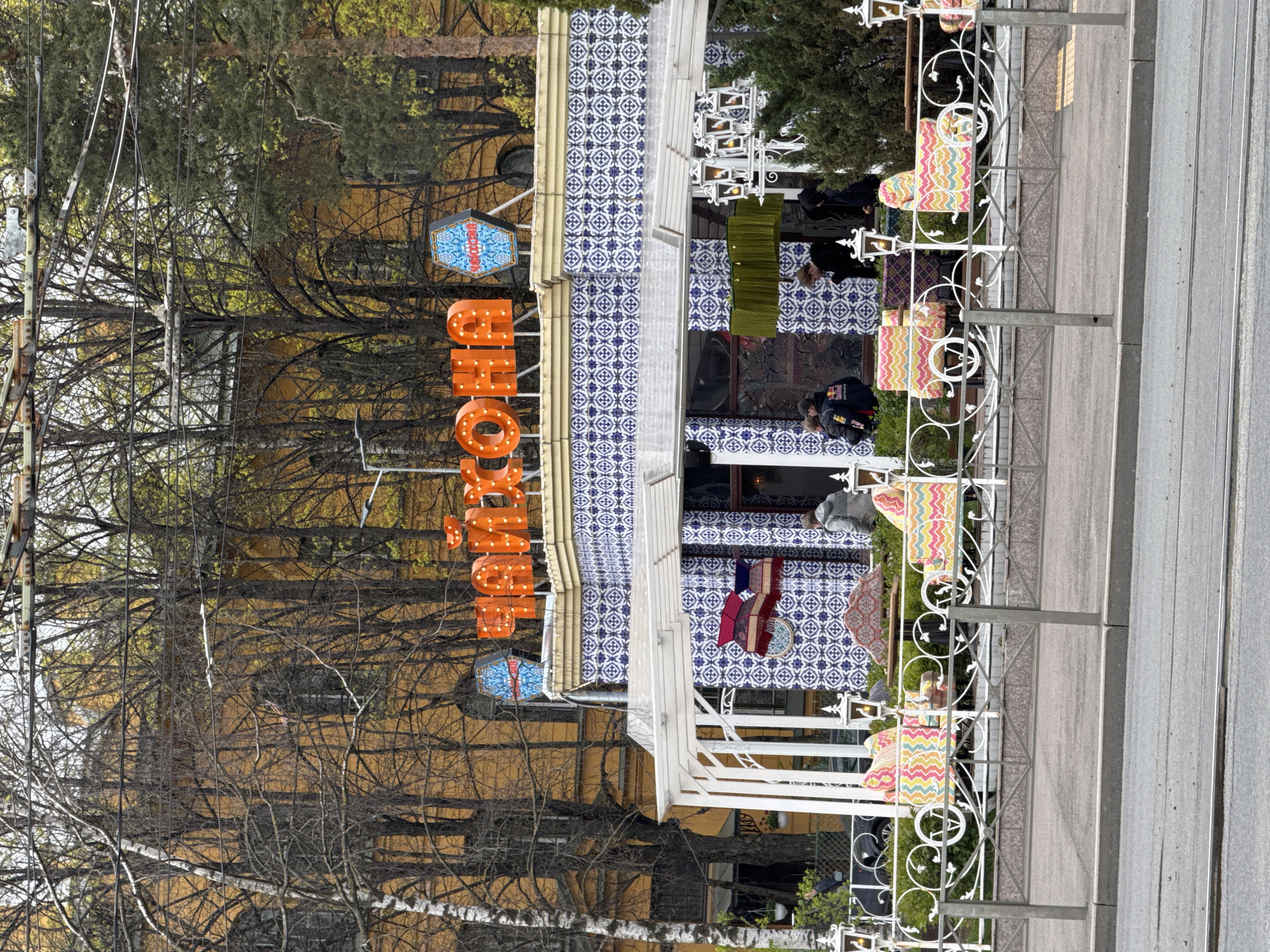 The image shows the exterior of a restaurant named "CHOKHO" (ХОЧУ) in Saint Petersburg, Russia. The restaurant has blue and white patterned tiles on its facade, and the name is displayed in large orange illuminated letters. There are people seated at tables outside the restaurant, and a decorative fence separates the seating area from the street. Trees are visible in the background. The scene appears to be daytime.