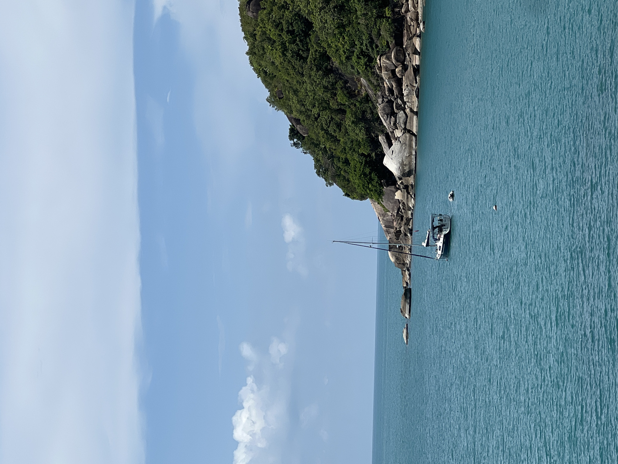 A sailboat is anchored in a clear blue body of water, close to a lush green island covered in trees and large rocks. The sky above is a light blue with scattered white clouds. The scene appears to be a coastal area in Baan Chaweng Noi, Thailand.