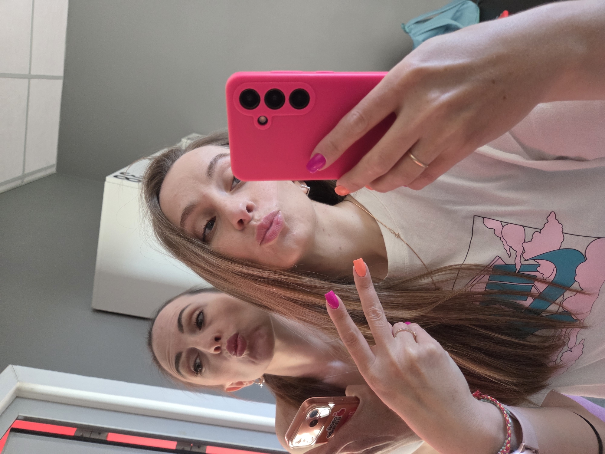A woman is taking a selfie in a mirror, possibly in a room with grey walls and white cabinets. She is holding a pink phone case with three camera lenses and is wearing a white t-shirt with a pink and blue design. Her nails are painted alternately with pink and orange colors. She is making a kissing face. In the mirror, her reflection is also making a kissing face and holding a phone with a clear case. The reflections gesture and appearance suggests it is a selfie taken in Russia. The location in the city of Birobidzhan, Russia, cannot be confirmed from this image alone.