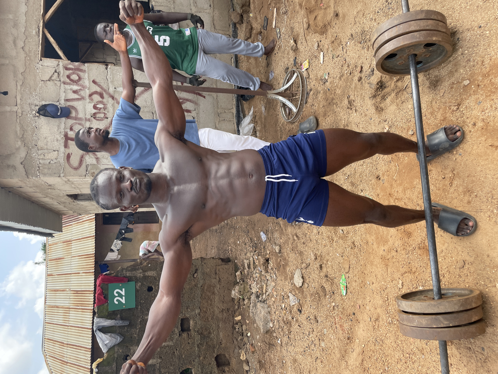 A shirtless, muscular man in blue shorts stands outdoors in Akwanga, Nigeria. He has arms raised, appearing to flex. To his right is a barbell with weights. Behind him, two men stand near a cinderblock building. Laundry hangs on a line. The ground is dirt with scattered debris. A sign with the number 22 is on the building. Another building with graffiti is in the background.
