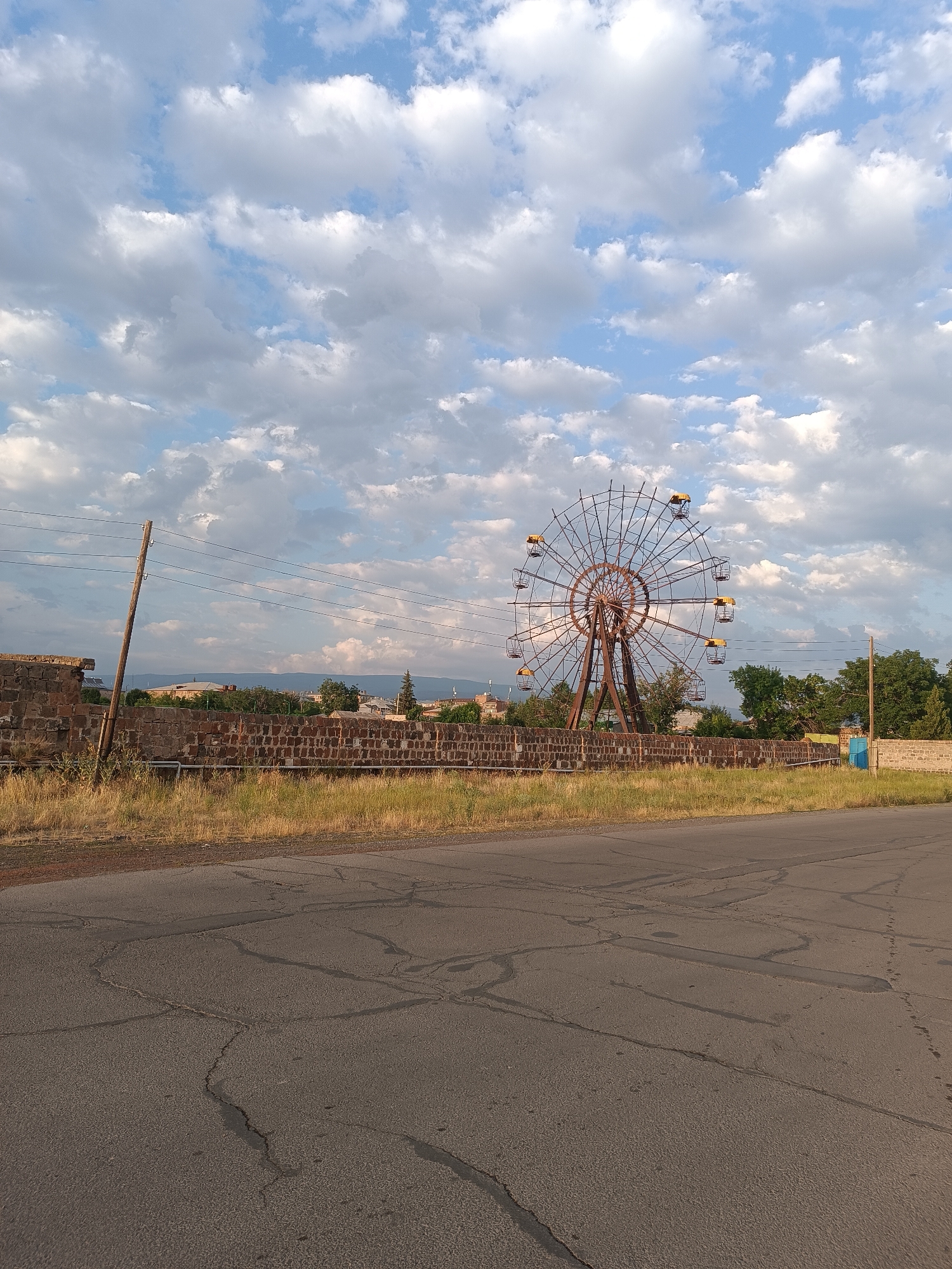 A slightly overcast sky with fluffy white clouds forms the backdrop for a rusty Ferris wheel, which appears to be disused. The Ferris wheel is located in Talin, Armenia. In the foreground, a cracked asphalt road stretches across the image, with dry, yellowed grass growing along its edge. A low, weathered brick wall runs horizontally behind the grass, and a wooden utility pole with multiple wires stands near the left side of the wall. Beyond the wall, trees and buildings can be seen in the distance, suggesting an urban or semi-urban setting. The overall scene evokes a sense of abandonment or a forgotten place.