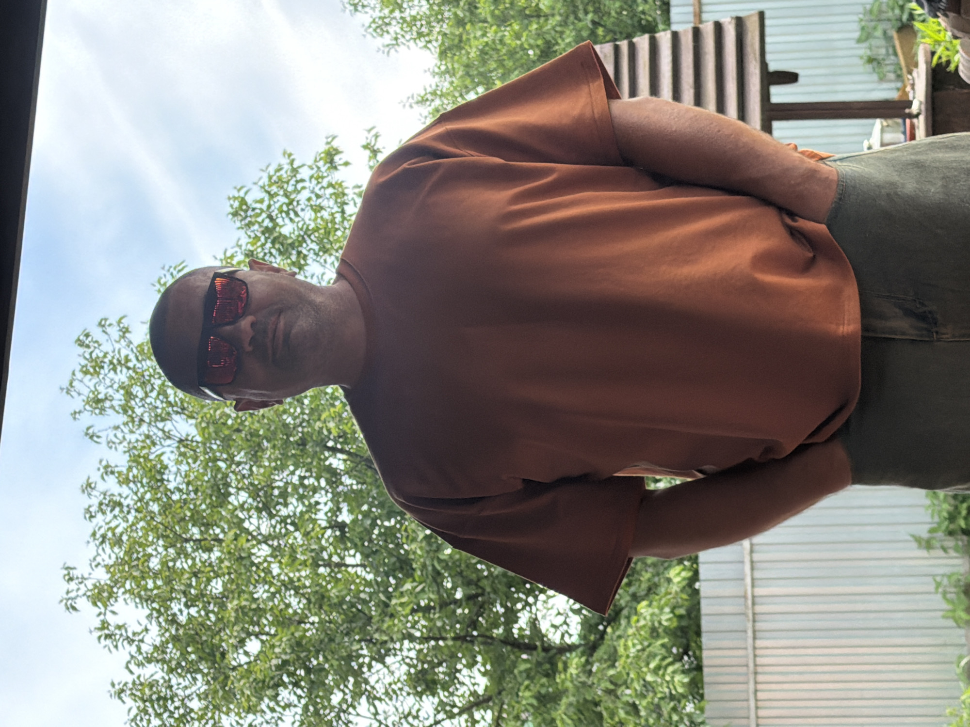 A portrait-oriented outdoor photograph captures a man looking directly at the camera. He has a shaved head and wears red-tinted, reflective sunglasses, a plain rust-colored or dark orange short-sleeved t-shirt, and olive green or gray trousers. His left arm is bent, with his hand resting near his hip.
The setting appears to be an outdoor area, possibly a garden or backyard, under a partly cloudy or overcast sky. Lush green foliage, likely from trees or bushes, dominates the left and upper background. To the right, a corrugated metal fence or structure is visible, suggesting an enclosure. The lighting indicates it is daytime. The man’s expression is neutral, with his eyes hidden by the sunglasses. There is no visible text in the image.