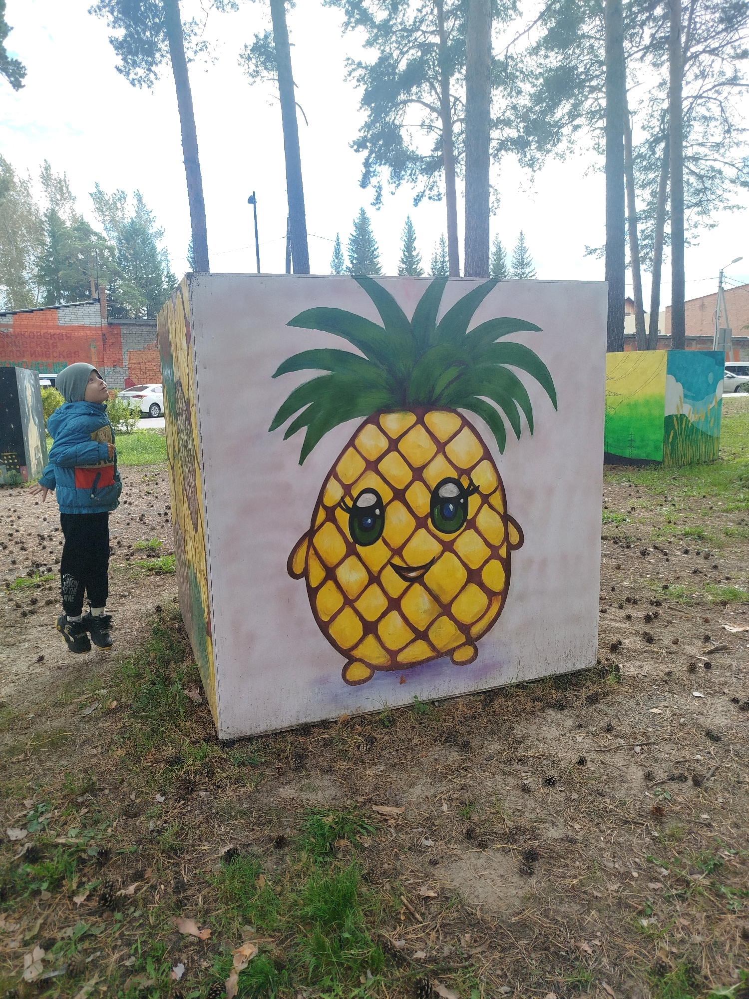 A young boy wearing a blue jacket, grey beanie, and black pants stands on a grassy area with pine cones scattered on the ground, looking up at a large concrete cube painted with a cartoonish pineapple. The pineapple has a smiling face with big, green eyes and a leafy crown. In the background, several other painted concrete cubes are visible, with one depicting a landscape with green hills and a blue sky. Pine trees tower over the scene, and a building with brickwork can be seen to the left. A white car is parked in the distance. The location is identified as Zavodoukovsk, Russia.