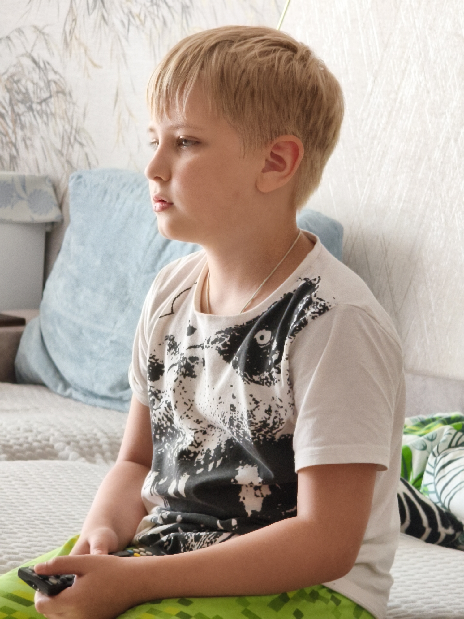 A fair-skinned boy with short blond hair is shown in profile, seated on a bed with a white textured wallpaper background. He is wearing a white t-shirt with a black abstract design. The boy is holding a black remote control in his right hand and looking to his left. He has a light blue cushion behind him and a green patterned blanket on his lap and to his right. The overall scene appears to be indoors, likely in a residential setting, and there is no indication of the specific location in Chaikovsky, Russia.