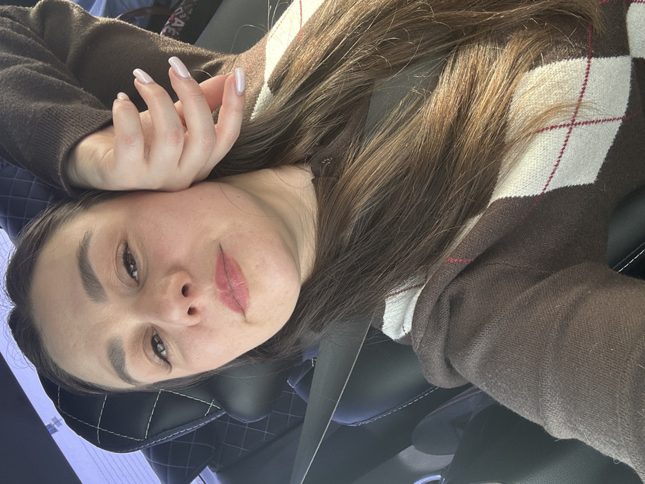 The image displays a single female individual inside the cabin of a vehicle. The specific location in the city of Mostovaya, Russia, cannot be determined from the visual information provided.
The subject has long, dark brown hair, centrally parted, cascading over her right shoulder and chest. Her complexion is light, with light pink lipstick on her lips and defined eyebrows. Her left hand, featuring manicured nails with an opaque, light pink or nude polish, is positioned against her left cheek and jawline. She wears a brown long-sleeved sweater adorned with a white and dark red argyle pattern across the chest and right arm. A black seatbelt is visible diagonally across her chest and left shoulder. The individual is looking directly towards the camera, suggesting a self-portrait.
The immediate background consists of a black car seat with a quilted pattern on the headrest, indicating a vehicle interior. A diffused light source, consistent with daylight, illuminates the scene from the right side, with a faint view of a rear window behind the subject's head.