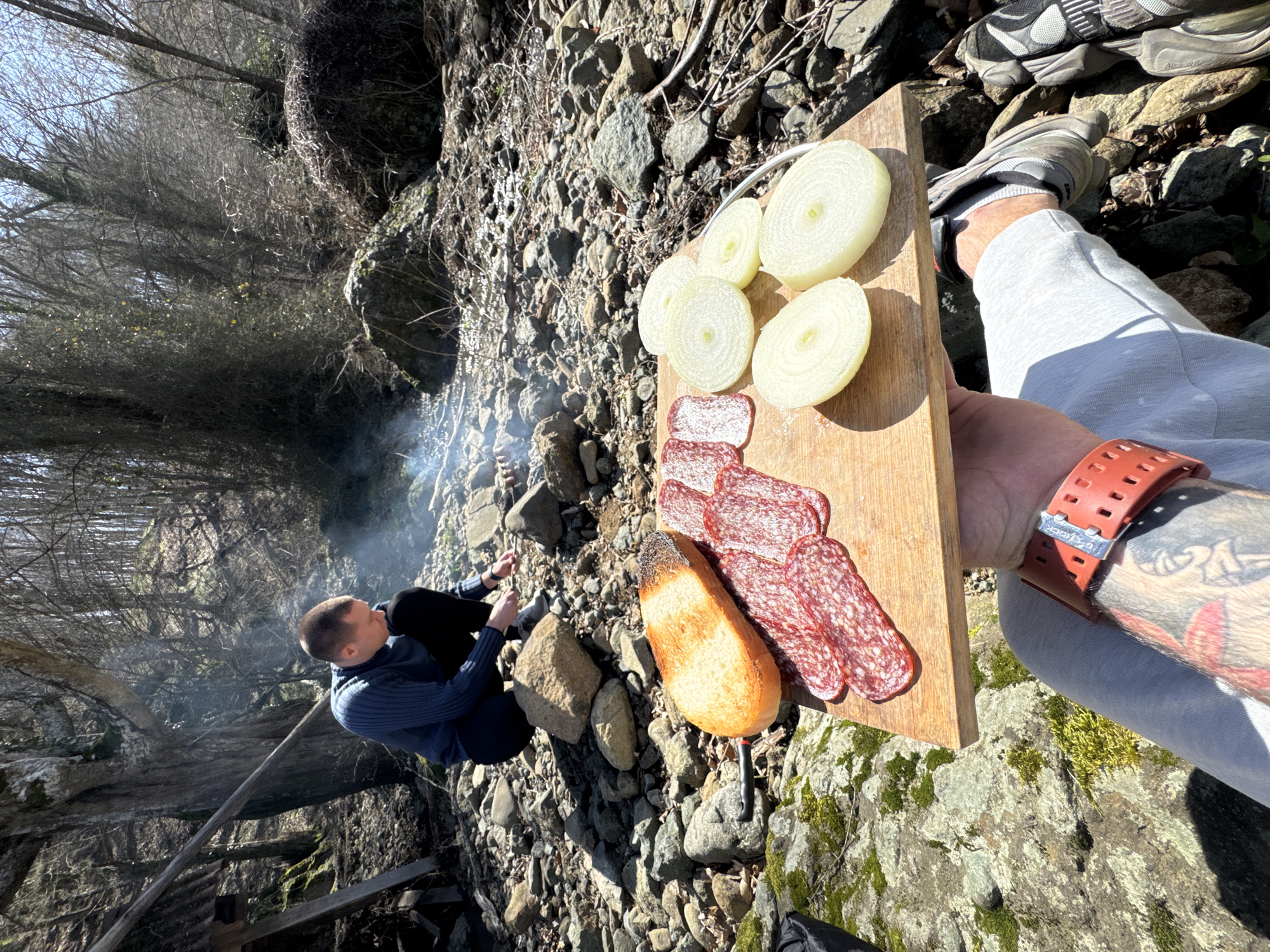 The image depicts an outdoor cooking and eating scene in a rocky, forested area, specifically identified as Fazanovo, Bulgaria. In the foreground, a person's tattooed forearm and lower leg are visible, holding a wooden cutting board. The forearm wears a red digital wristwatch. On the cutting board, there is a toasted piece of bread, several slices of reddish salami or sausage, and five thick slices of white onion. The individual's leg is dressed in light grey sweatpants and grey sneakers with white laces, resting on a moss-covered rock surface.
In the mid-ground, a male individual is crouched over a small, smoldering fire, identified by a plume of light smoke rising from a cluster of rocks. He is wearing a dark blue long-sleeved ribbed sweater and dark pants. The ground surrounding him is composed of numerous grey and brown rocks and pebbles, characteristic of a dry riverbed or forest floor. To the left, a partial, rudimentary structure made of wooden beams and corrugated metal is visible. The background consists of a dense forest of deciduous trees with bare branches, suggesting late winter or early spring. Sunlight illuminates the scene from the right, casting shadows and indicating clear weather.