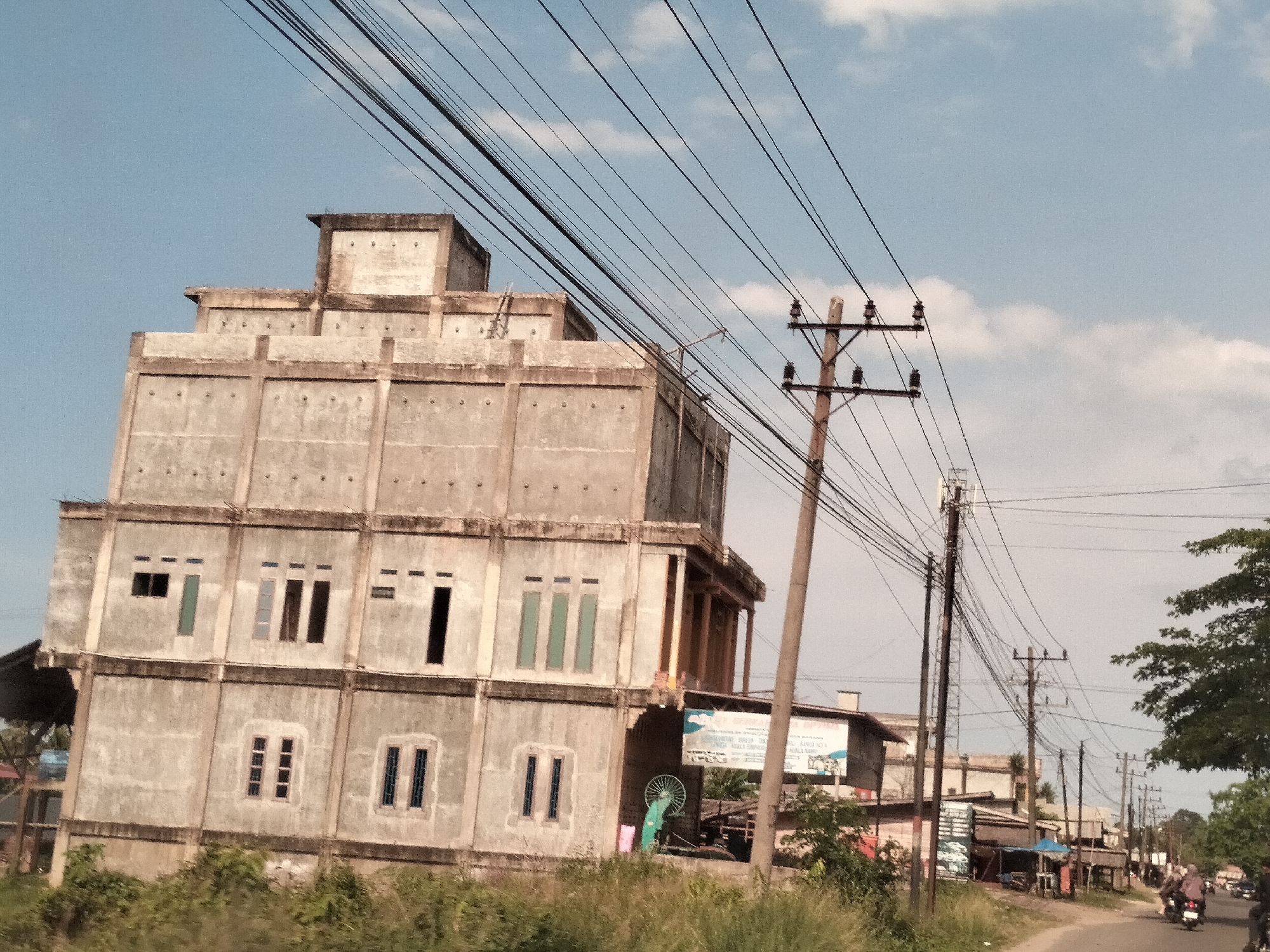 The image shows an unfinished concrete building in Teupinbatee, Indonesia. The building has four levels, with a tiered structure on top. There are several windows on the lower levels. Power lines are strung across the sky and a wooden utility pole stands near the building. The road is visible with a few motorcycles. There are additional buildings and stalls visible along the road. Overgrown vegetation is present in the foreground. The sky is light blue with some clouds.