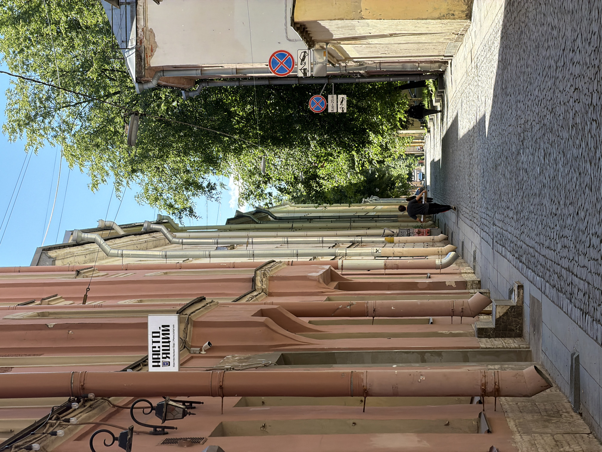 This is a medium shot, taken from a low angle, of a narrow cobblestone street in Saint Petersburg, Russia, on a sunny day. The street is lined with tall, narrow buildings with pastel-colored facades, predominantly pink and beige. The buildings have ornate architectural details, including arched doorways and protruding drainpipes. On the left side of the street, a white sign with Cyrillic text and a logo is mounted on the pink building. Further down the street, two people are walking away from the camera, one carrying a large flat object. Another person is walking further ahead on the right side of the street. Traffic signs indicating parking restrictions are visible on the right side of the street, near a yellow building. Lush green trees line the right side of the street, casting shadows on the cobblestones. The sky is a clear, bright blue.