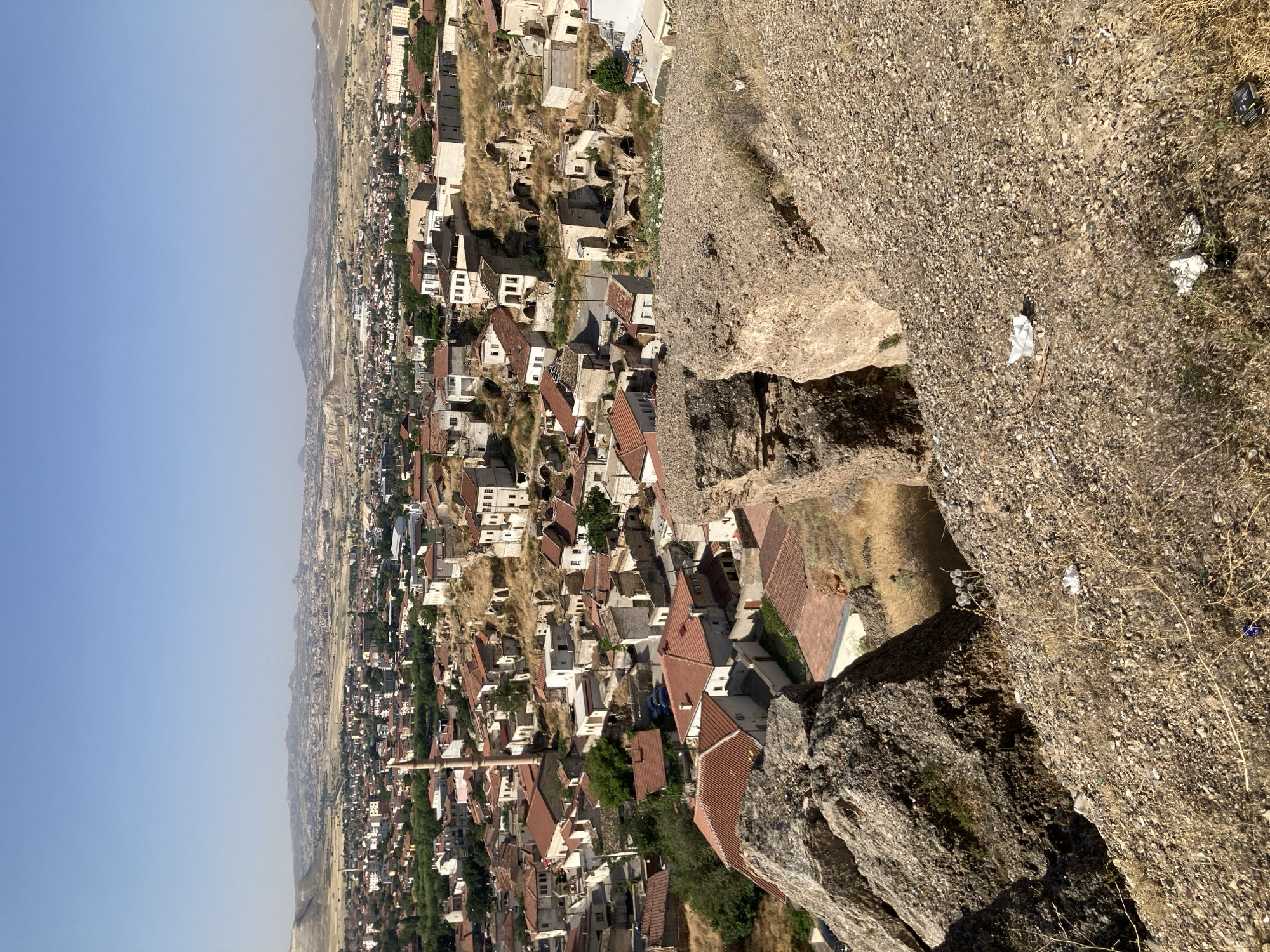The image depicts a panoramic view of Avanos İlçe Merkezi, Turkey, on a clear, sunny day. In the foreground, large rock formations covered in gravel and dry vegetation dominate the lower right and left portions of the frame. A crevice is visible between these rocks. The middle ground reveals a dense collection of buildings with red-tiled roofs and stone or stucco walls, characteristic of the region's architecture. Several structures appear to be ancient, with some showing signs of disrepair and integration into the rocky landscape. A tall, slender minaret rises from the center of the town. Beyond the immediate cluster of buildings, the town extends into the distance, with more buildings spread across a gently undulating, arid landscape. In the far background, rolling hills and mountains form the horizon under a pale blue sky. The environment appears dry and somewhat parched, with sparse vegetation visible throughout the scene, particularly around the buildings and on the hillsides. No people are visible in the image. The overall impression is of a historic settlement nestled within a rugged, natural landscape.