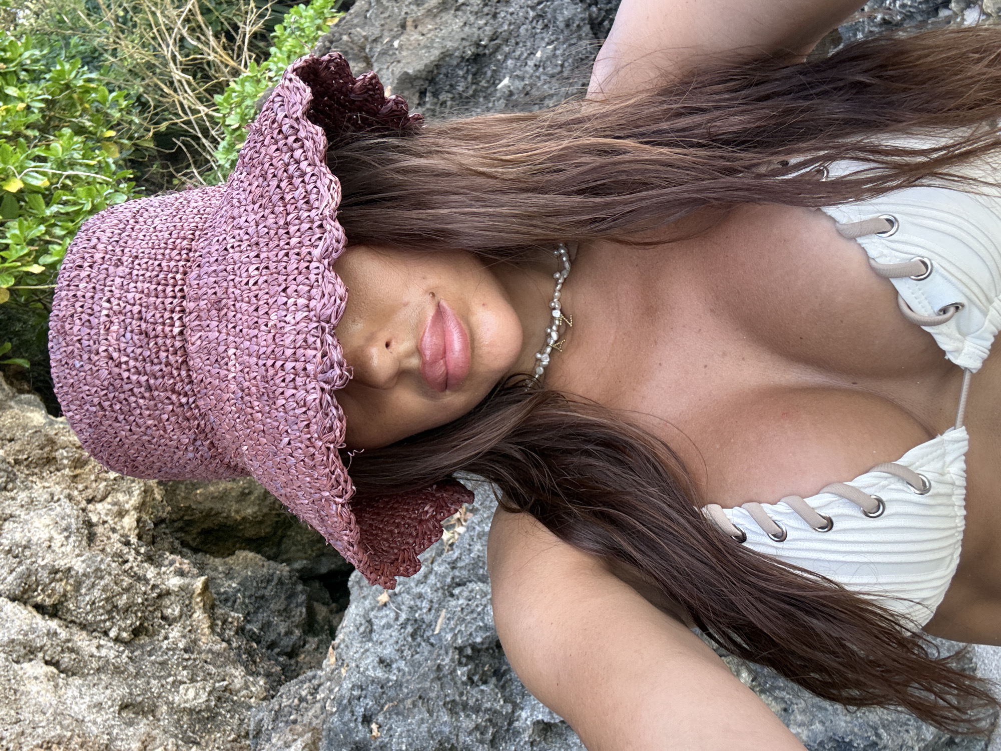 A woman is reclining on a rocky surface in Kuta Selatan, Indonesia. She is wearing a white bikini top with lace-up detailing and a pink straw hat that obscures part of her face. Her long brown hair is spread out around her. She is also wearing a pearl necklace. The background features rocks and vegetation.