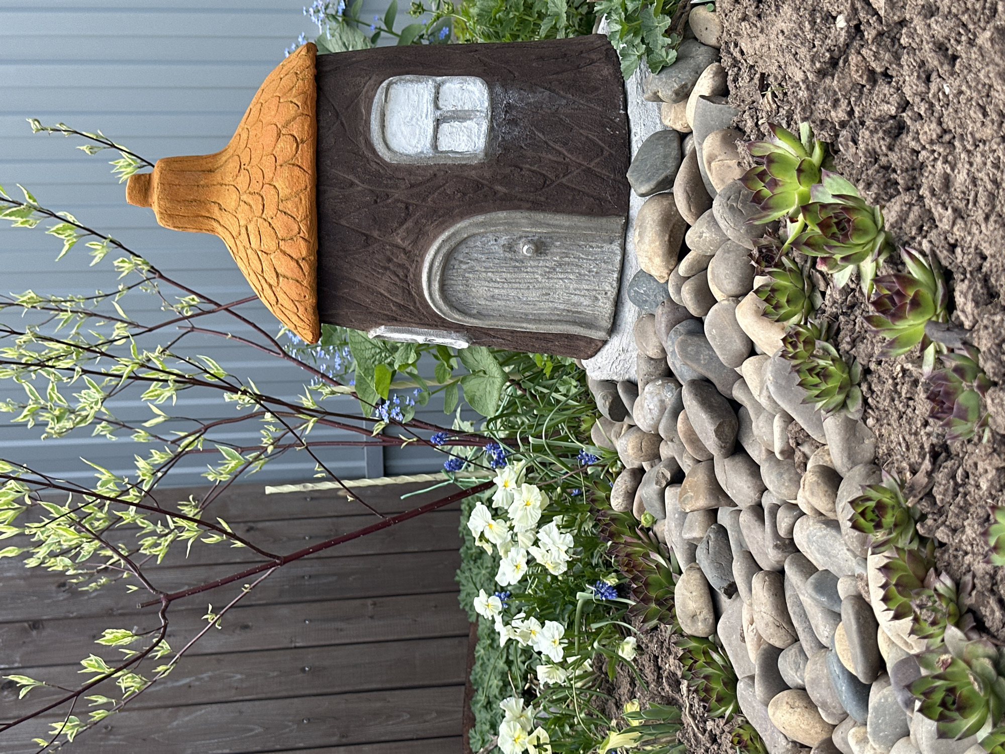The image shows a miniature fairy house situated in a garden setting. The house is made of brown material with a textured wood-like appearance and has an orange, textured roof. A small, white-framed window and a gray arched door with a knob are visible on the house.
The house is surrounded by various plants, including white and blue flowering plants, and succulents. A border of gray river stones lines the edge of the garden bed. To the left of the house, a tree branch with small variegated leaves extends into the frame. A brown wooden fence is partially visible in the lower left corner of the image.
The location in Ludorvai, Russia, is not verifiable from the image.