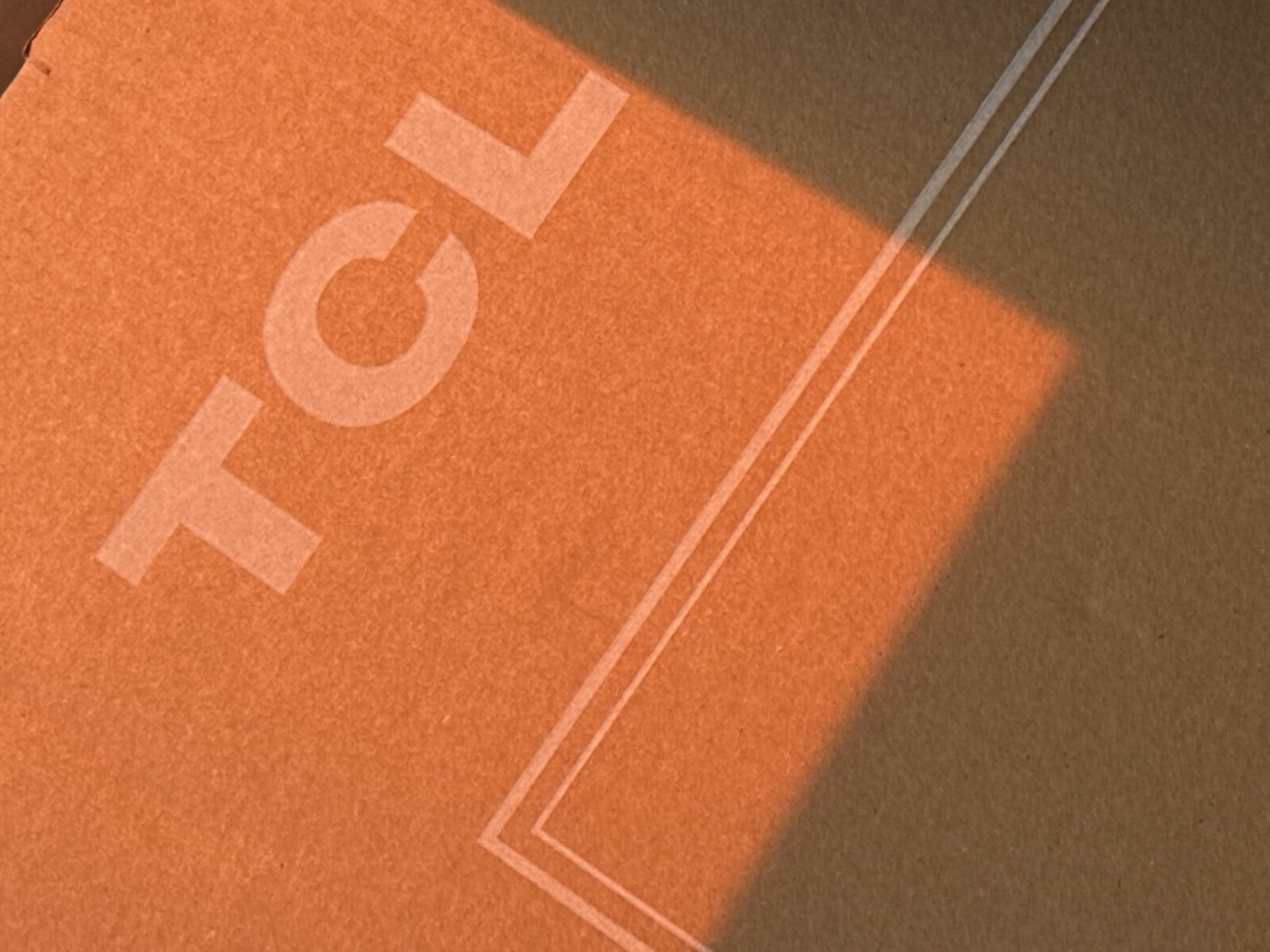 The image shows a section of a cardboard box with the letters "TCL" printed in white. There are two parallel white lines forming a corner. The lighting is uneven, with a shadow cast across the right side of the box. The cardboard appears to be a standard brown color. The location is not discernible from the provided image.