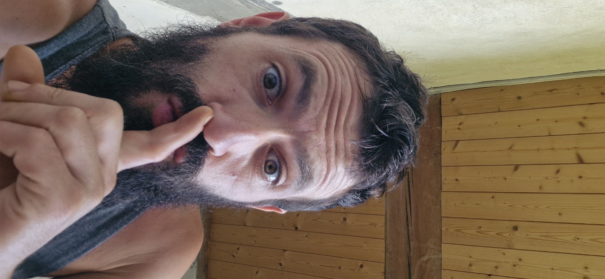 A man with a dark beard and messy hair is looking at the camera with his finger pressed to his nose, as if to silence someone. He is wearing a gray tank top. The background appears to be an outdoor setting with a wall and a wooden ceiling. The man's facial expression is one of surprise or jest.