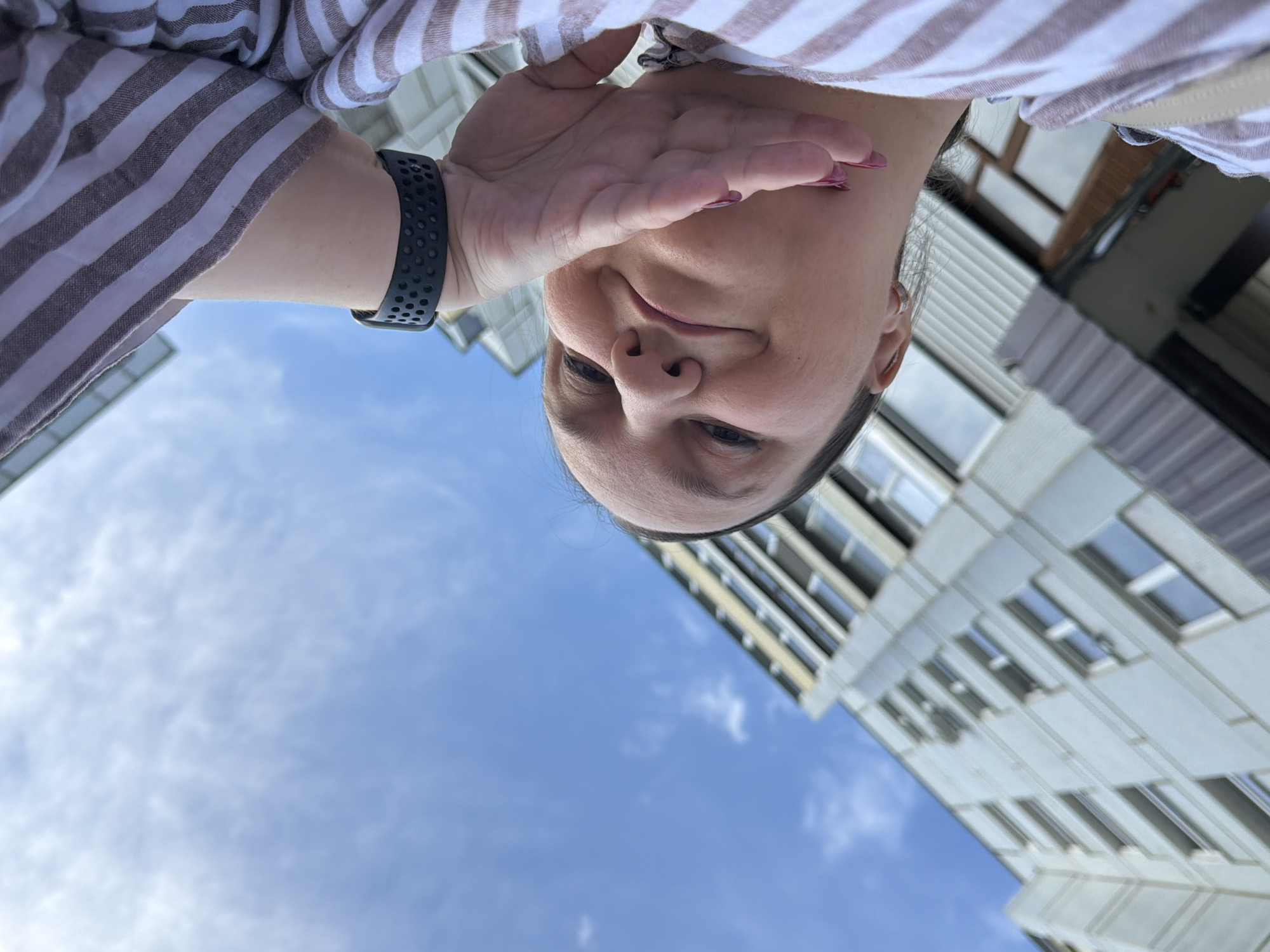 In a downward-facing selfie, a person wearing a horizontally striped brown and white long-sleeved shirt and a black smartwatch on their left wrist covers their face with their right hand. The person's head is angled, with their eyes and nose visible. They appear to be outdoors, with a bright blue sky and scattered clouds in the background, along with a portion of a modern building with windows. The image was taken in Chaikovsky, Russia, on a clear day.