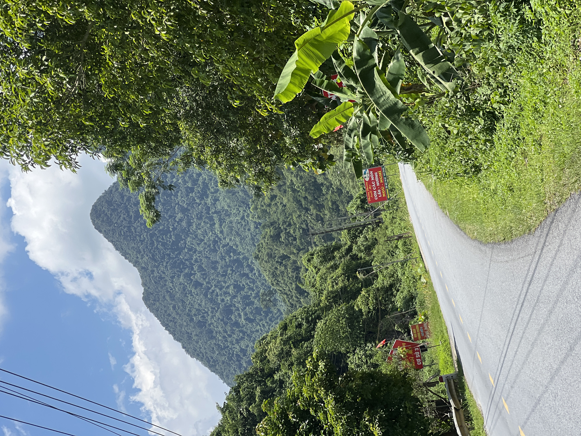 The image depicts a winding asphalt road in a rural area, likely in Thuận Hòa Commune, Vietnam, given the signage. The road curves to the right, bordered by lush green vegetation, including banana trees and dense undergrowth. On the left side of the road, a metal guardrail is visible, and beyond it, a steep, verdant mountain rises prominently against a bright blue sky with scattered white clouds. Several red signs are posted along the side of the road, some featuring Vietnamese text and graphics. Overhead, power lines crisscross the sky. The overall scene conveys a sense of natural beauty and tranquility, characteristic of a countryside landscape.