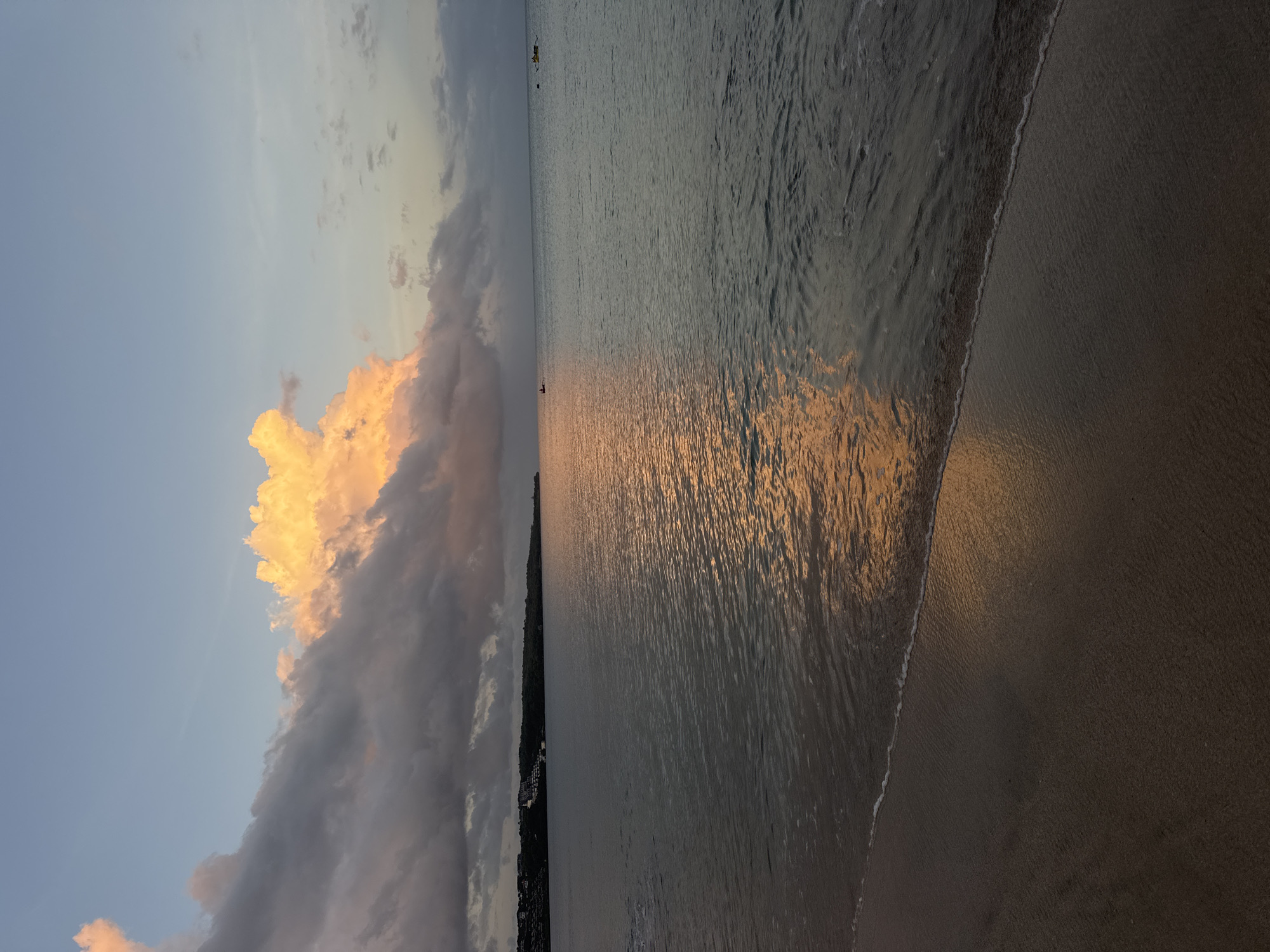 The image captures a dusk scene at Kuta Beach, Indonesia. The sky is partially covered by clouds, with some illuminated by the setting sun, casting a golden glow. The ocean reflects the colors of the sky, creating a shimmering effect on the water's surface. A coastline with dense vegetation and buildings is visible. A beach with waves is located on the bottom right. There are a few boats in the distance.