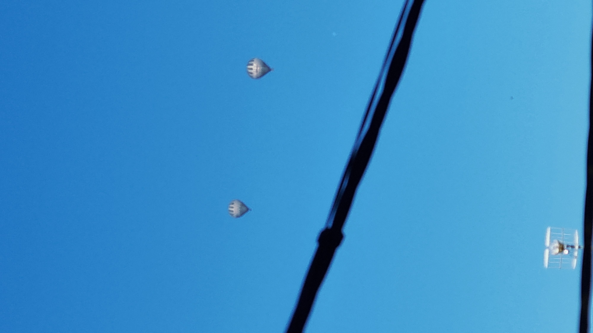 Two hot air balloons are visible in a clear blue sky. A dark, thick cable runs diagonally across the lower half of the image. In the bottom left corner, a miniature replica of a biplane is seen. The scene appears to be outdoors with good visibility. The location is specified as Sant Joan Les Fonts, Spain.