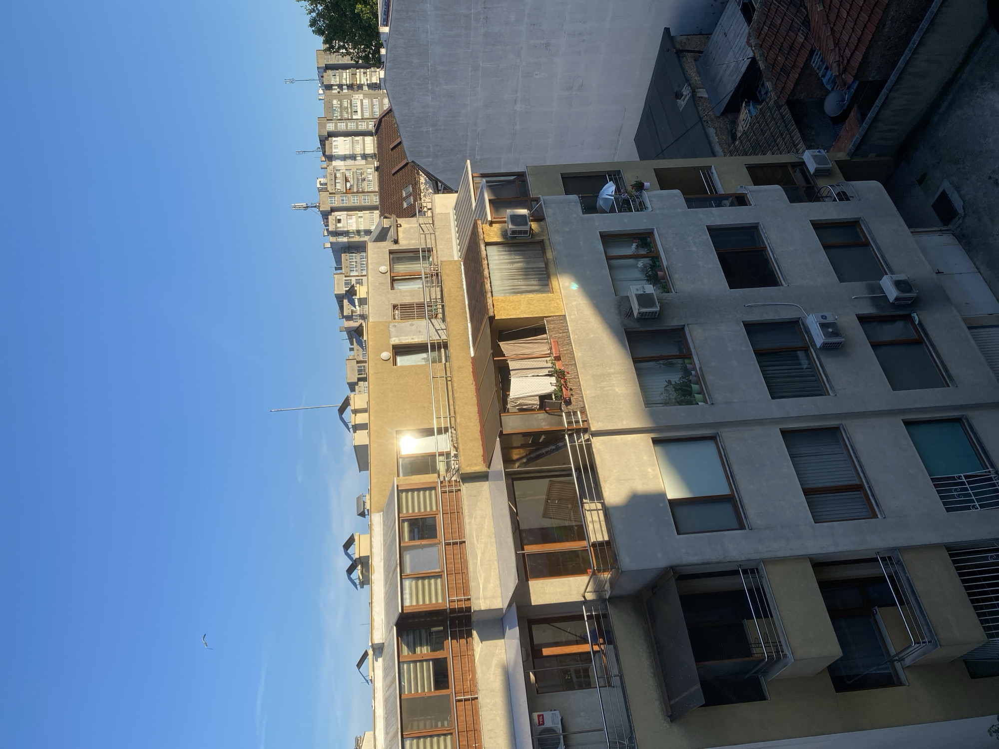 The image captures a view of apartment buildings in Varna, Bulgaria, under a clear blue sky. The buildings exhibit a mix of architectural styles and colors, with some featuring balconies and window units for air conditioning. The sun casts shadows across the facades, indicating it is likely morning or late afternoon. In the distance, a row of taller, more uniform apartment blocks suggests a more developed urban area. A single bird is visible flying in the upper left portion of the frame, adding a touch of nature to the cityscape. The foreground shows the upper floors of several residential buildings with visible windows, balconies, and external air conditioning units. The overall impression is of a typical urban residential environment in Varna.