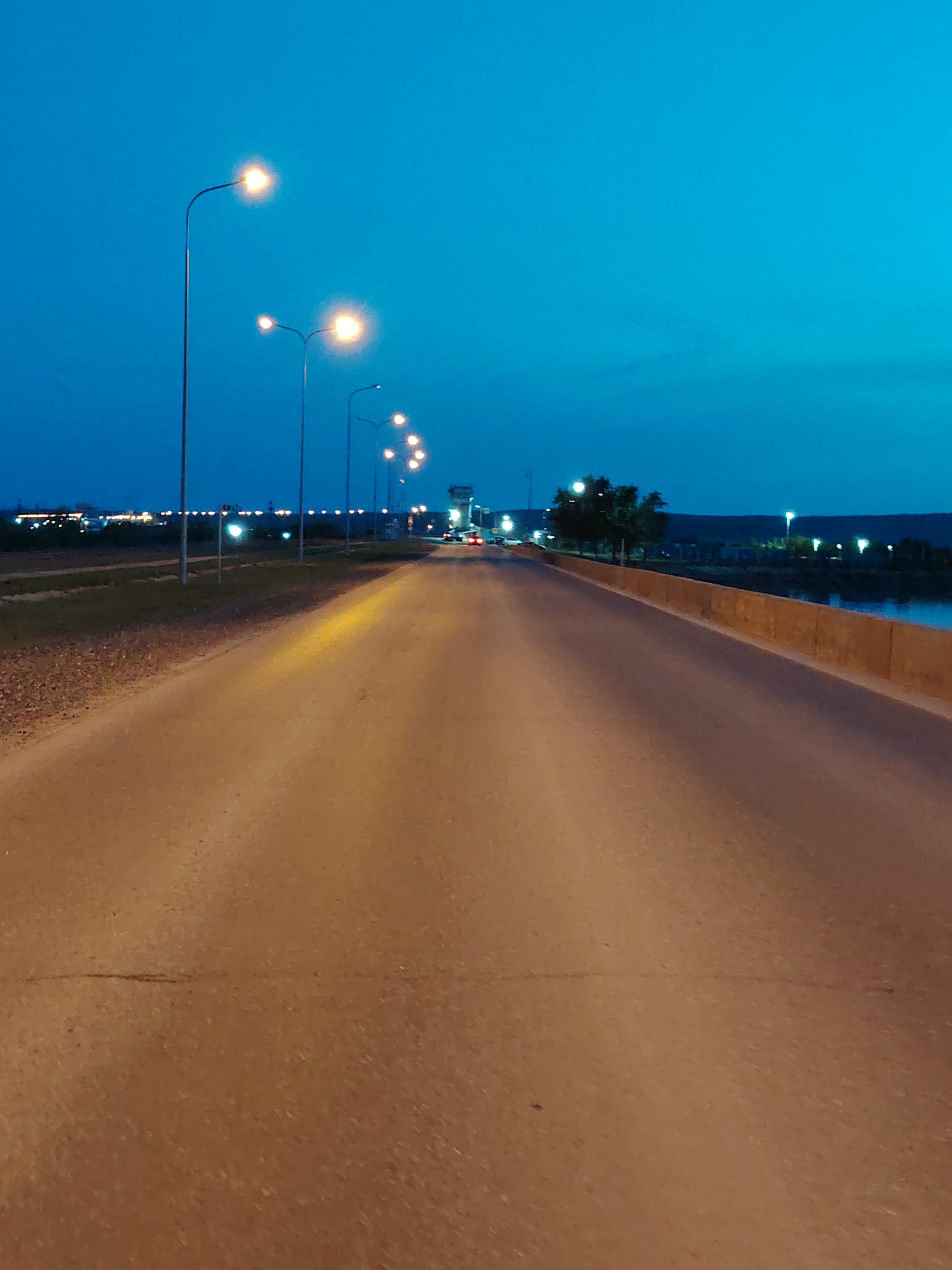 The image shows a straight, empty road at dusk, illuminated by a row of streetlights on the left side. The road surface is a tan color. On the left side of the road is a gravel shoulder and a low grassy area. Beyond that, there are more lights visible in the distance. On the right side of the road is a low concrete barrier and beyond that appears to be water with lights reflecting on the surface. Trees are sparsely present along the right side of the road. A control tower is visible farther down the road, with a car's headlights also visible. The sky is a deep blue color. The location is likely a road on the outskirts of Chaikovsky, Russia, possibly leading towards an industrial area near the Kama River.