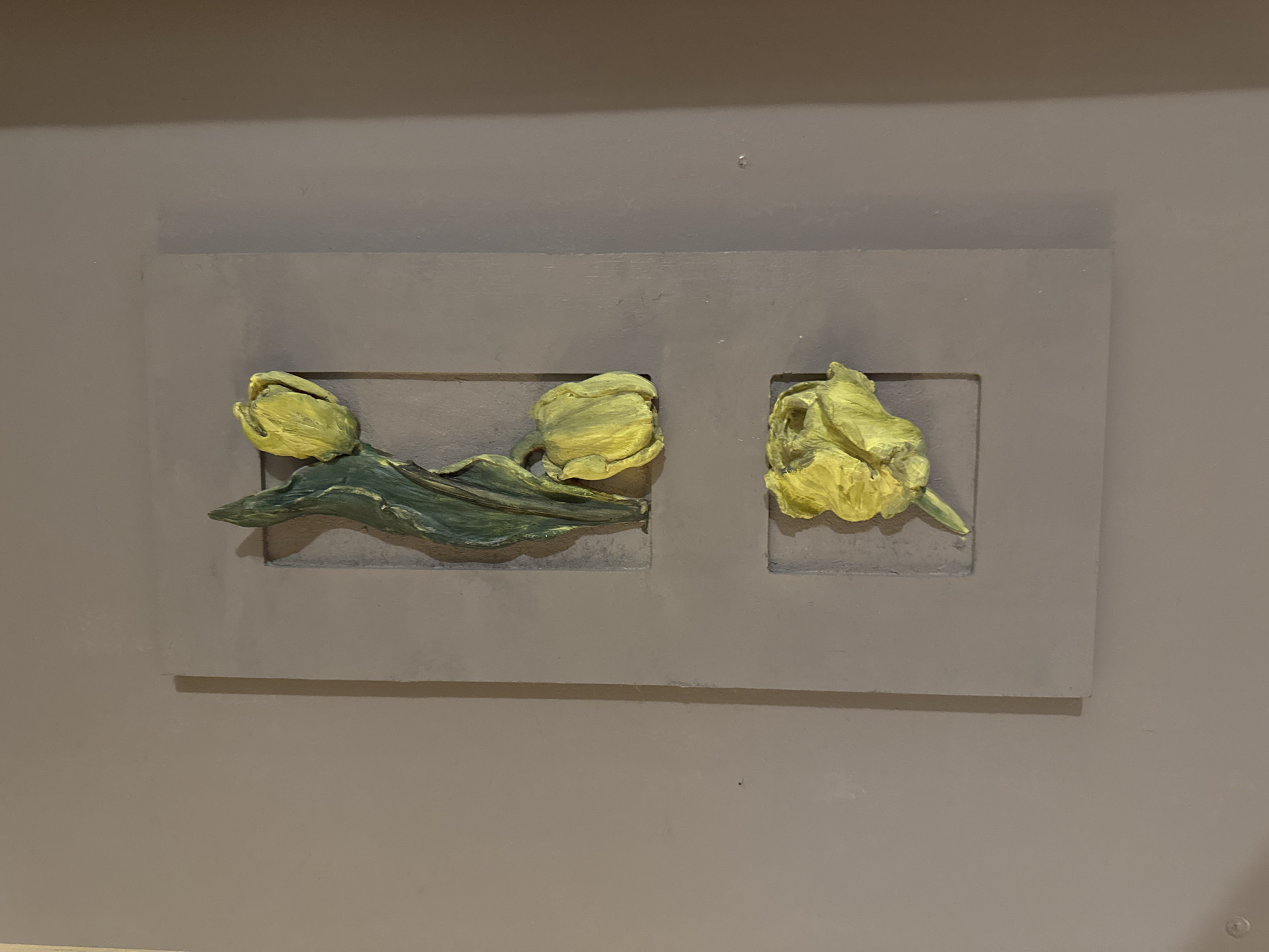 This media file displays a rectangular relief sculpture mounted on a flat, neutral-colored wall. The sculpture features three stylized yellow tulip blooms with green leaves and stems. The tulips are arranged in two recessed square panels within the larger rectangular frame. The left panel contains two blooms and a cluster of leaves, with one bloom slightly obscuring another. The right panel displays a single tulip bloom. The sculpting technique gives the flowers a textured, almost painterly appearance, with visible brushstrokes and varying shades of yellow and green. The overall impression is one of a decorative wall art piece. There is no visible text in the image, nor any specific cues to time of day, weather, or location beyond the indoor setting suggested by the wall. The absence of people or any indication of an event suggests the image is a static depiction of the artwork itself.