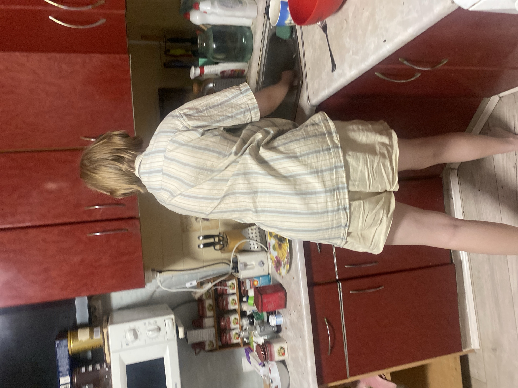 The image depicts a young person standing at a kitchen sink in a dimly lit kitchen, likely in Chornomorsk, Ukraine. The person, viewed from the back, has blond hair and is wearing a light-colored, striped short-sleeved shirt and light-colored shorts. They are engaged in washing dishes, with their right hand in the sink. The kitchen features dark red cabinets, a white countertop, and various kitchen items, including bottles, jars, a microwave, and a toaster. The overall impression is one of a casual, lived-in domestic space.