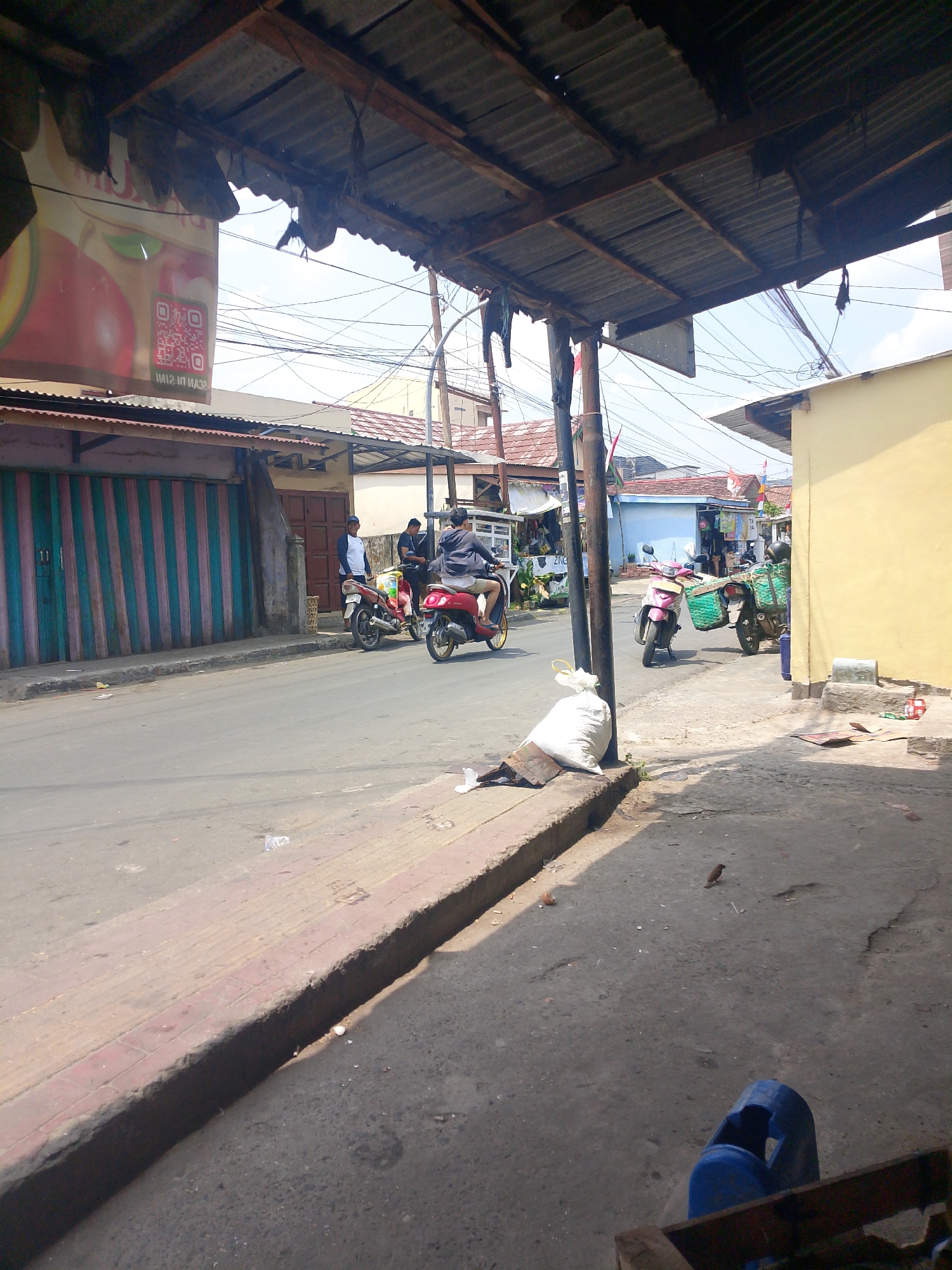 The image depicts a street scene in Palembang, Indonesia, likely in a commercial or residential area with a mixture of buildings and makeshift structures. The foreground features a textured concrete sidewalk with a raised curb, adjacent to a paved road. A white sack of what appears to be waste is placed on the edge of the sidewalk, with a small bird nearby. A blue plastic bin is visible in the lower right corner.
The midground shows a street with several motorcycles, indicating ongoing activity. One motorcycle, a red scooter, is ridden by a person wearing a dark jacket and light-colored pants, moving away from the camera. Another motorcycle, also a red scooter, is parked on the side of the road. Further down the street, a pink scooter with a green basket is parked, with a person in dark clothing standing next to it. Two other individuals are near a parked motorcycle. The buildings lining the street have varied architectural styles, with some featuring corrugated metal roofs and roll-up doors painted with vertical stripes of green, pink, and white. A yellow building is prominent on the right side of the street.
The background reveals more buildings, overhead utility wires, and a cloudy sky. A large banner with a QR code and an image of an apple is visible hanging from a corrugated metal roof structure, suggesting a commercial establishment. The overall impression is that of a bustling, everyday street scene in a city, with evidence of commerce and daily life.