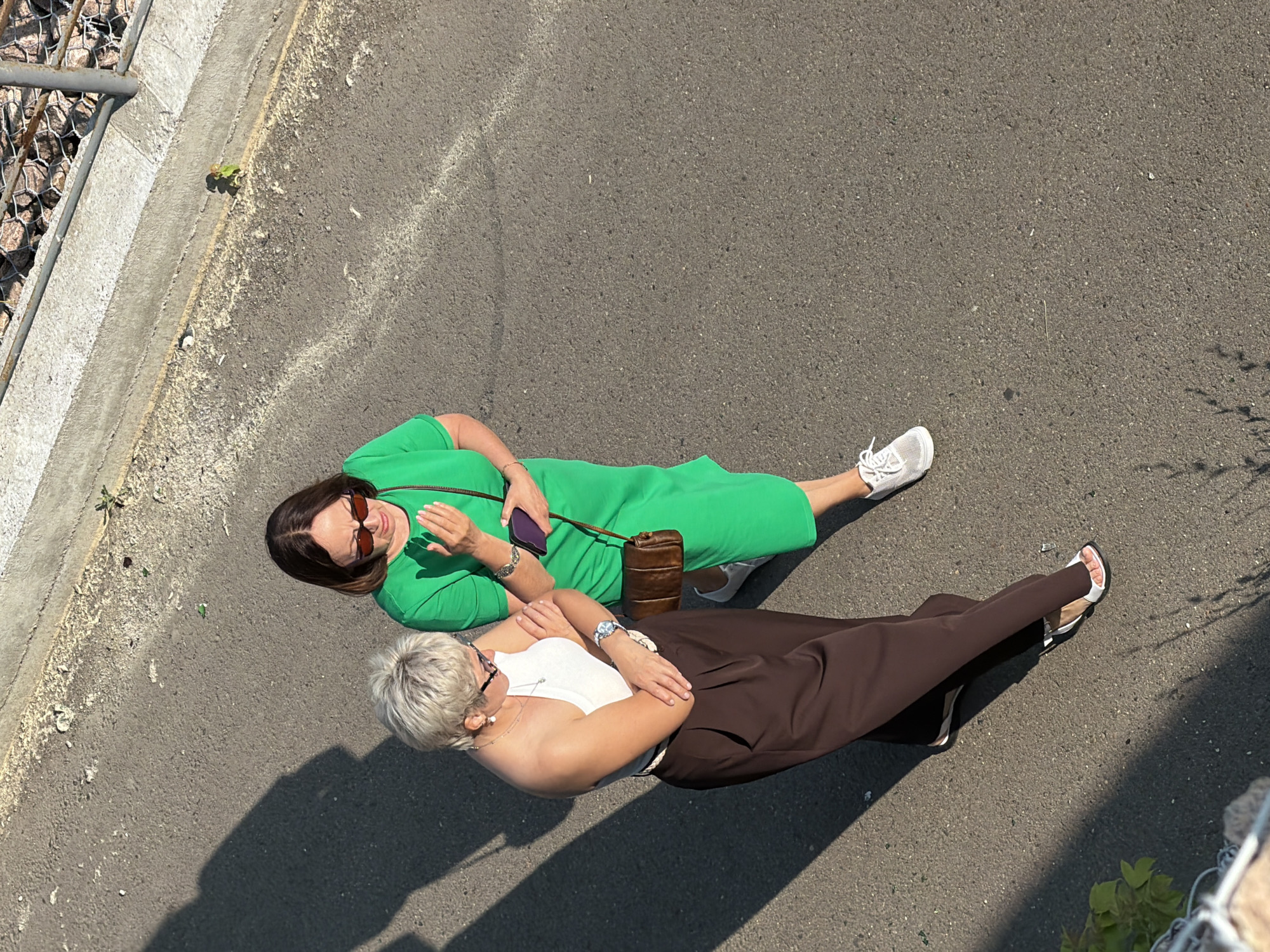 Two women are lying on their sides on a paved surface. The woman on the left is wearing a bright green dress and sunglasses. She is holding a dark smartphone in her left hand and has her right arm bent at the elbow, with her hand resting near her head. A brown, cylindrical bag is positioned behind her. She has dark hair and is looking downwards.
The woman on the right has short, light-colored hair and is wearing a white top and dark brown, wide-leg pants. She has on white sandals. Her arms are crossed in front of her chest, and she is also looking downwards. Both women are wearing sunglasses, suggesting it is a sunny day.
The scene appears to be outdoors, with a paved surface that has some faint white markings. A concrete barrier with wire mesh and rocks is visible along the left edge of the image. Green foliage can be seen in the lower right corner, casting a shadow. The lighting suggests it is daytime, with strong sunlight creating distinct shadows. The overall impression is one of relaxation or a posed moment in an outdoor urban or park setting.
